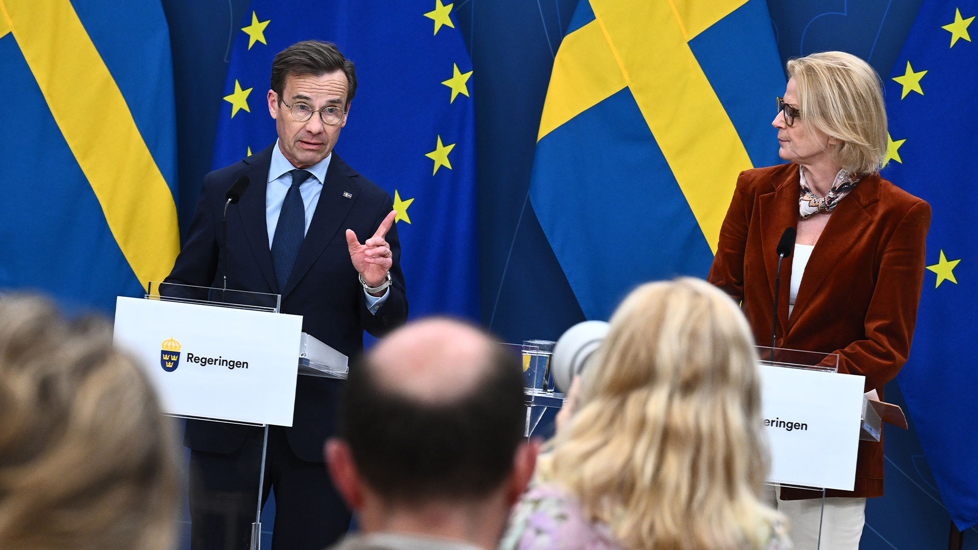 Två regeringsrepresentanter vid en presskonferens framför svenska och EU-flaggor.