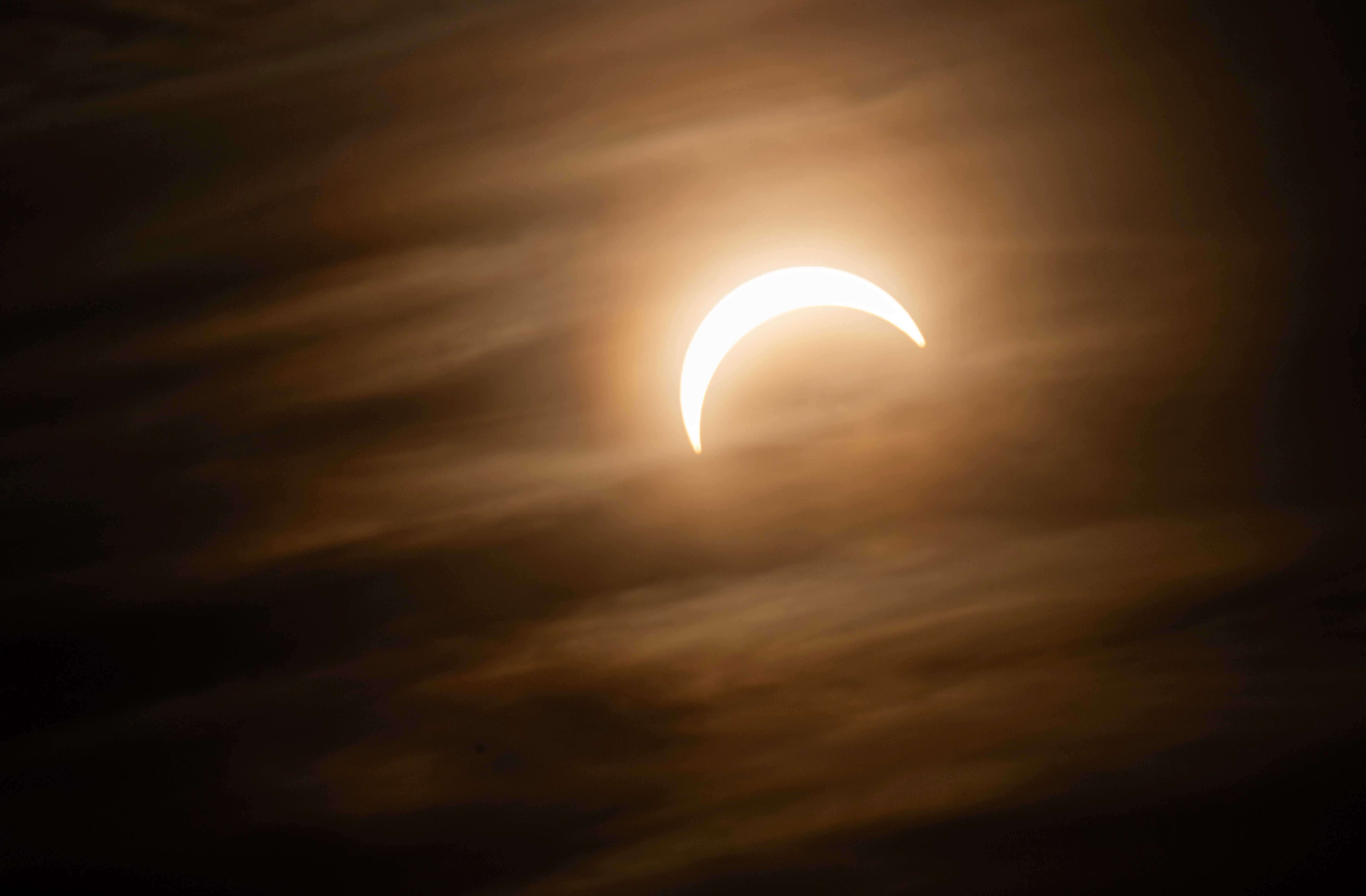 Partiell solförmörkelse där månen skymmer delen av solen, vilket skapar en ljus krescent mot en mörk molnig himmel.