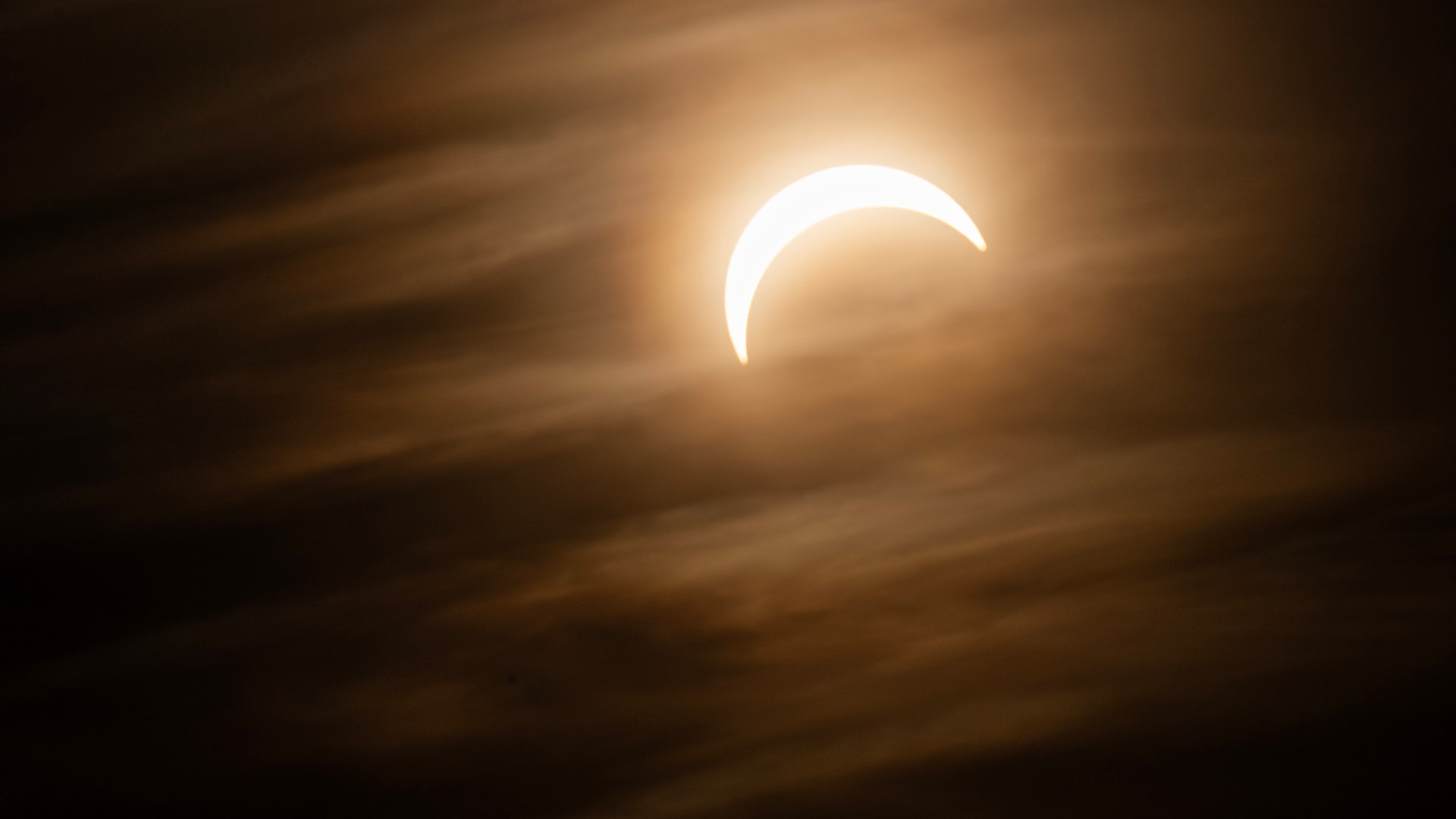 Partiell solförmörkelse där månen skymmer delen av solen, vilket skapar en ljus krescent mot en mörk molnig himmel.