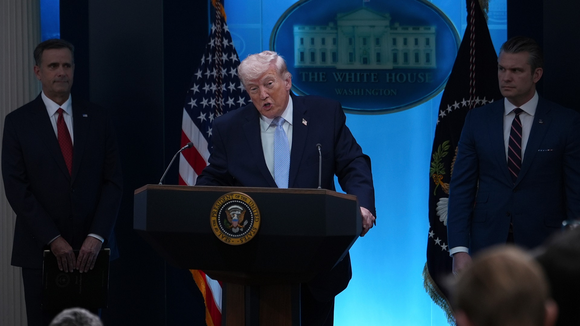 En man i mörk kostym talar vid ett podium med presidentens sigill framför en blå bakgrund med Vita huset-logotypen. Två män i kostym står på vardera sidan om honom.