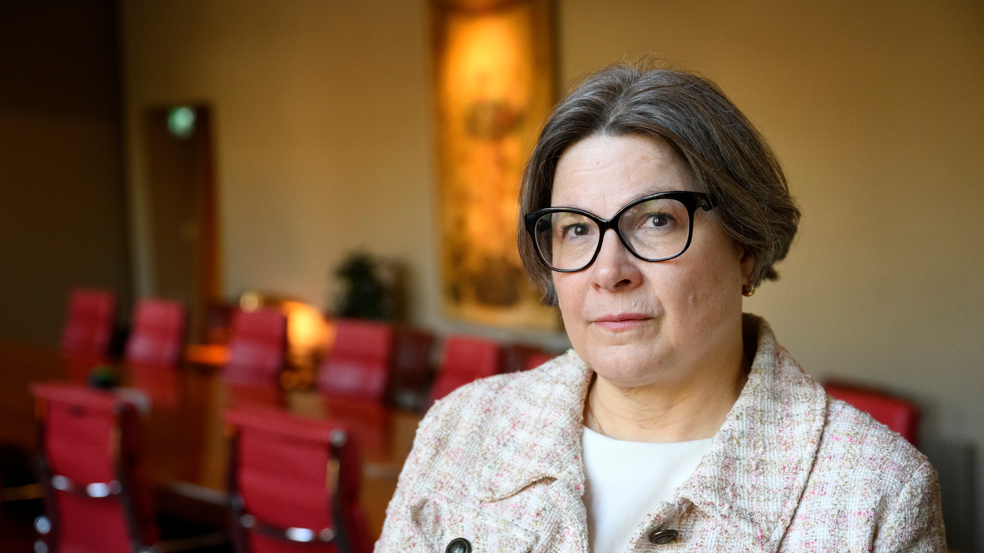 Kvinna med glasögon sitter i en auditorium med röda stolar i bakgrunden.