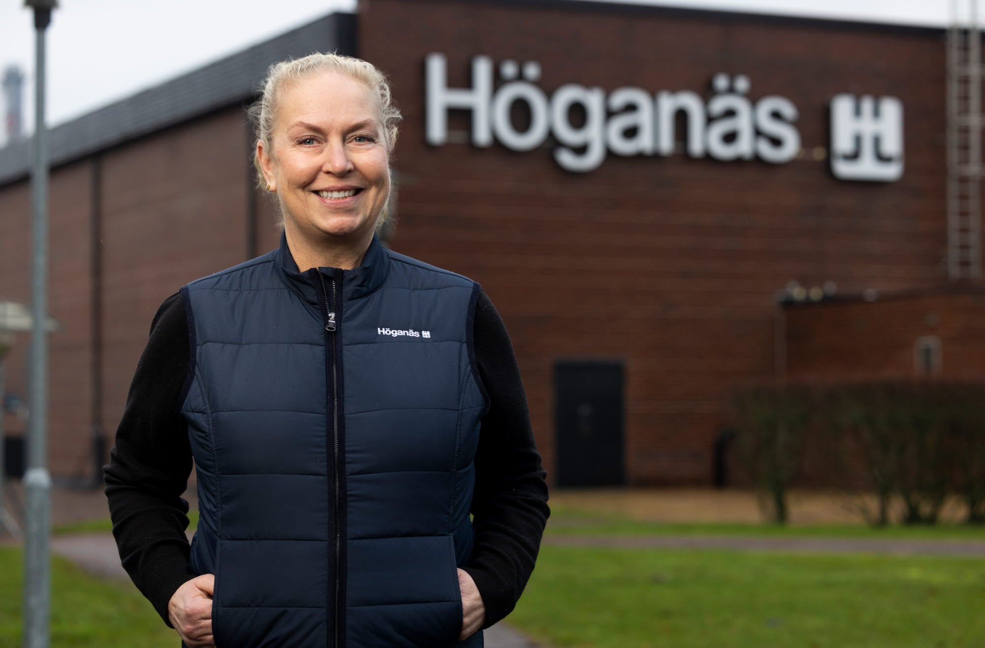 Porträtt av en kvinna som står framför en tegelbyggnad med Höganäs-skylt. Hon bär en mörkblå väst och svart långärmad skjorta.