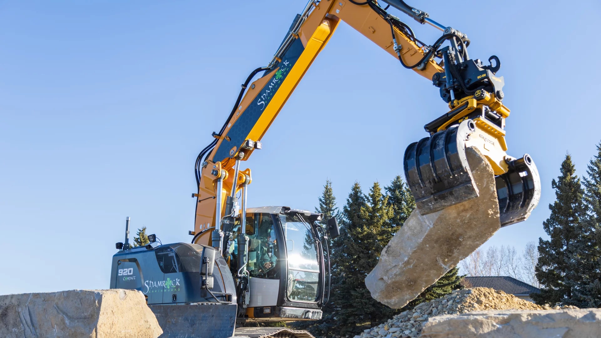 En gul grävmaskin med skopa arbetar med material på en byggarbetsplats mot en klar blå himmel.