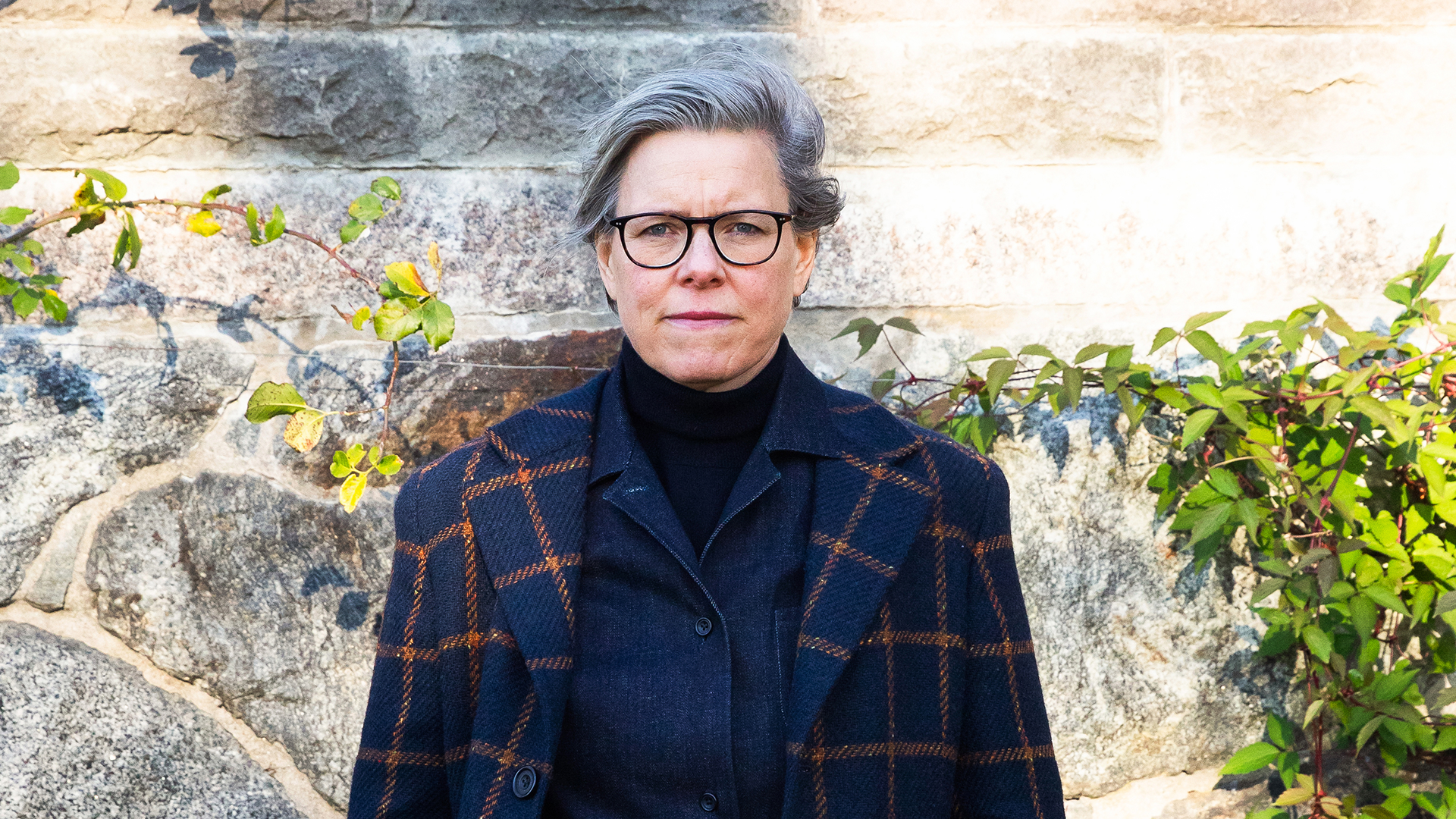 Författaren Lena Andersson står utomhus, framför en vägg av sten klädd i murgröna, och tittar in i kameran.