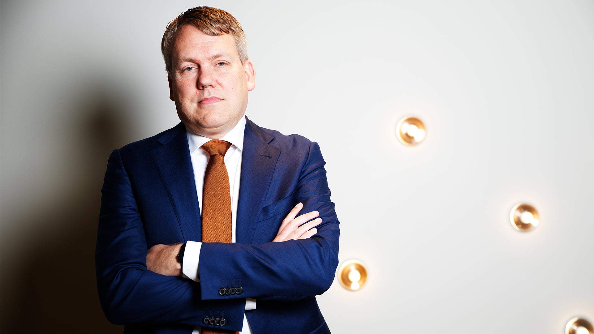 Porträtt av en man i mörkblå kostym och orange slips, med armarna i kors, mot en vit bakgrund med dekorativa guldiga element.