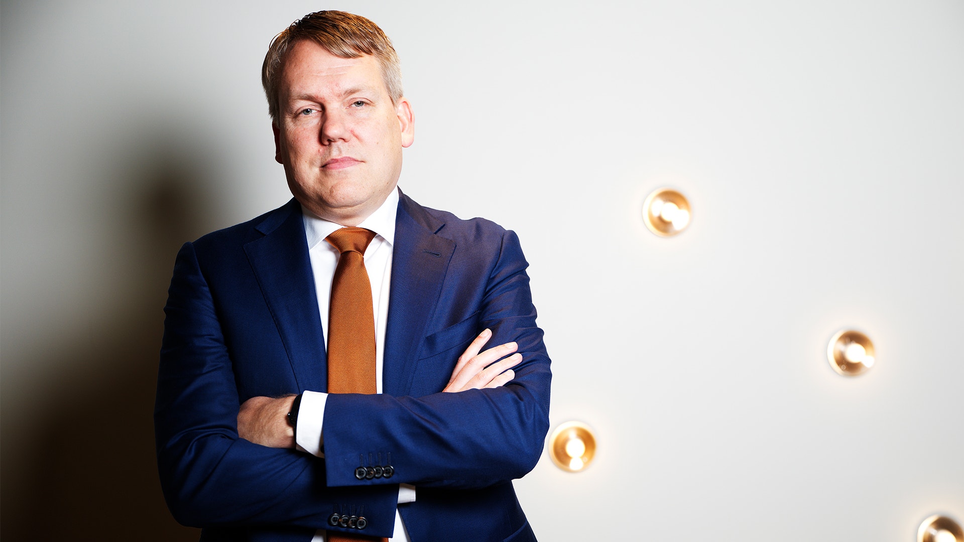 Porträtt av en man i mörkblå kostym och orange slips, med armarna i kors, mot en vit bakgrund med dekorativa guldiga element.