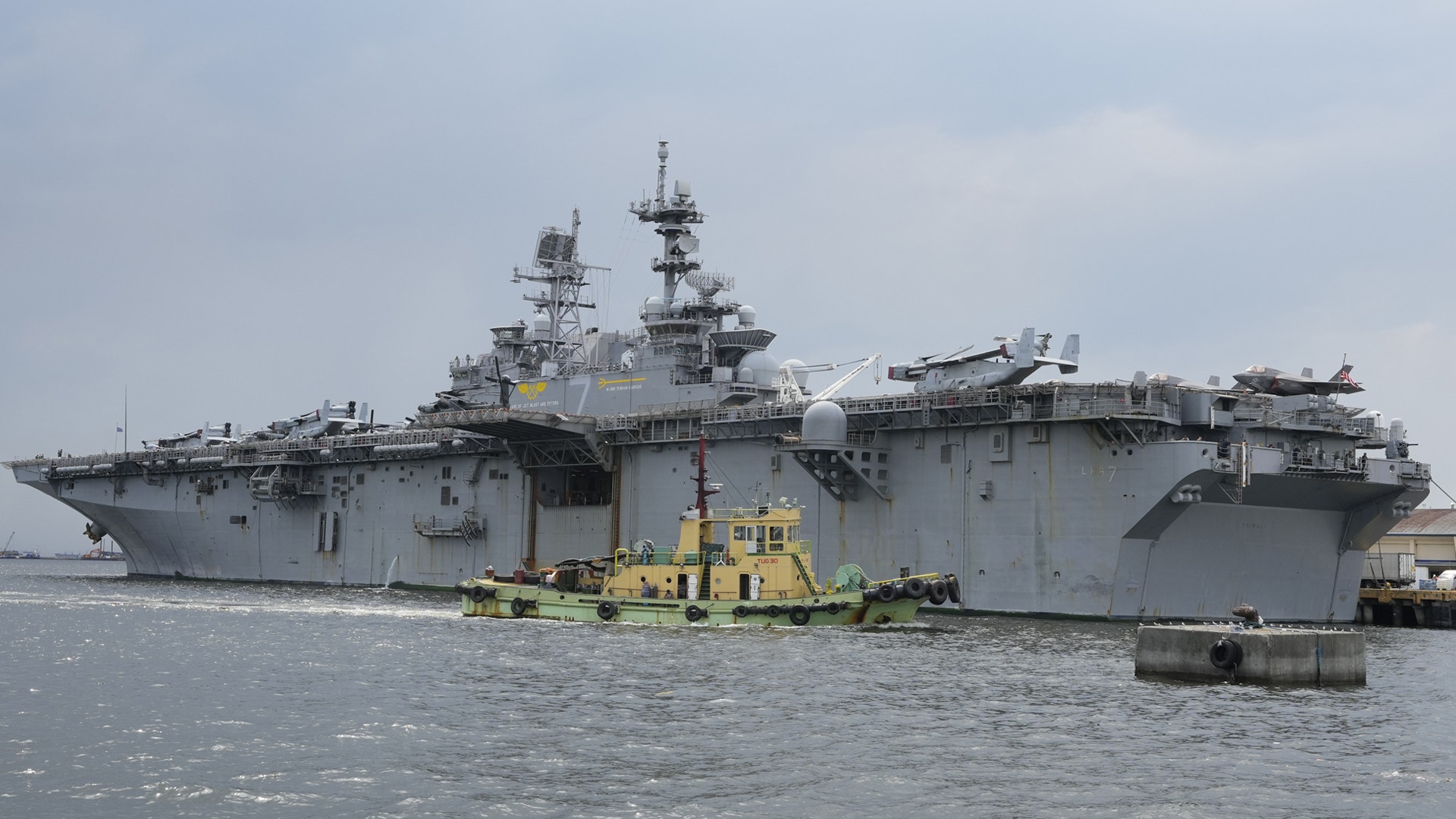 Stort militärt hangarfartyg i vatten med gult bogserbåt vid sidan
