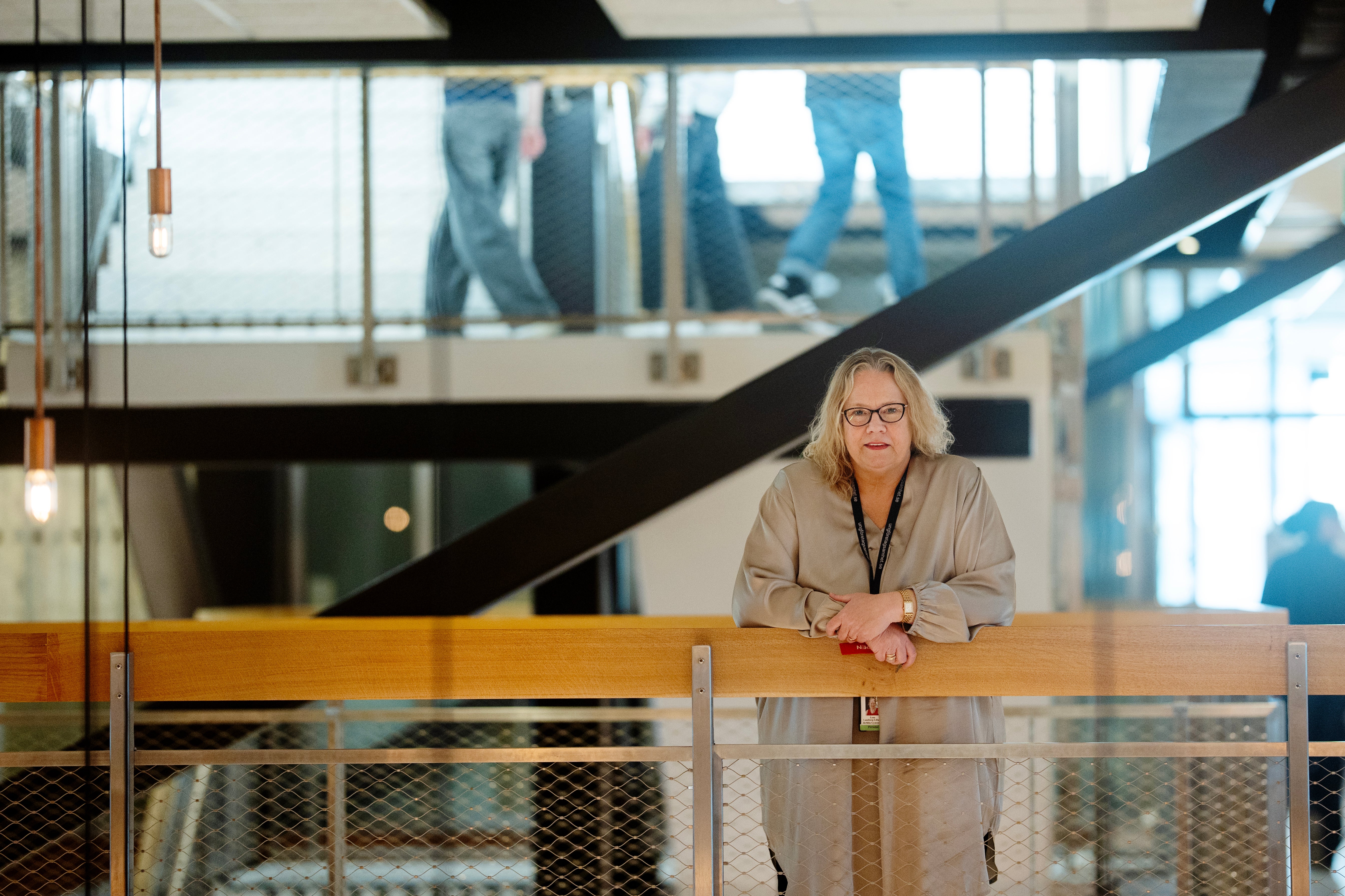 Porträtt av en kvinna med glasögon som står vid ett räcke i en modern byggnad med stora fönster och trappor i bakgrunden.
