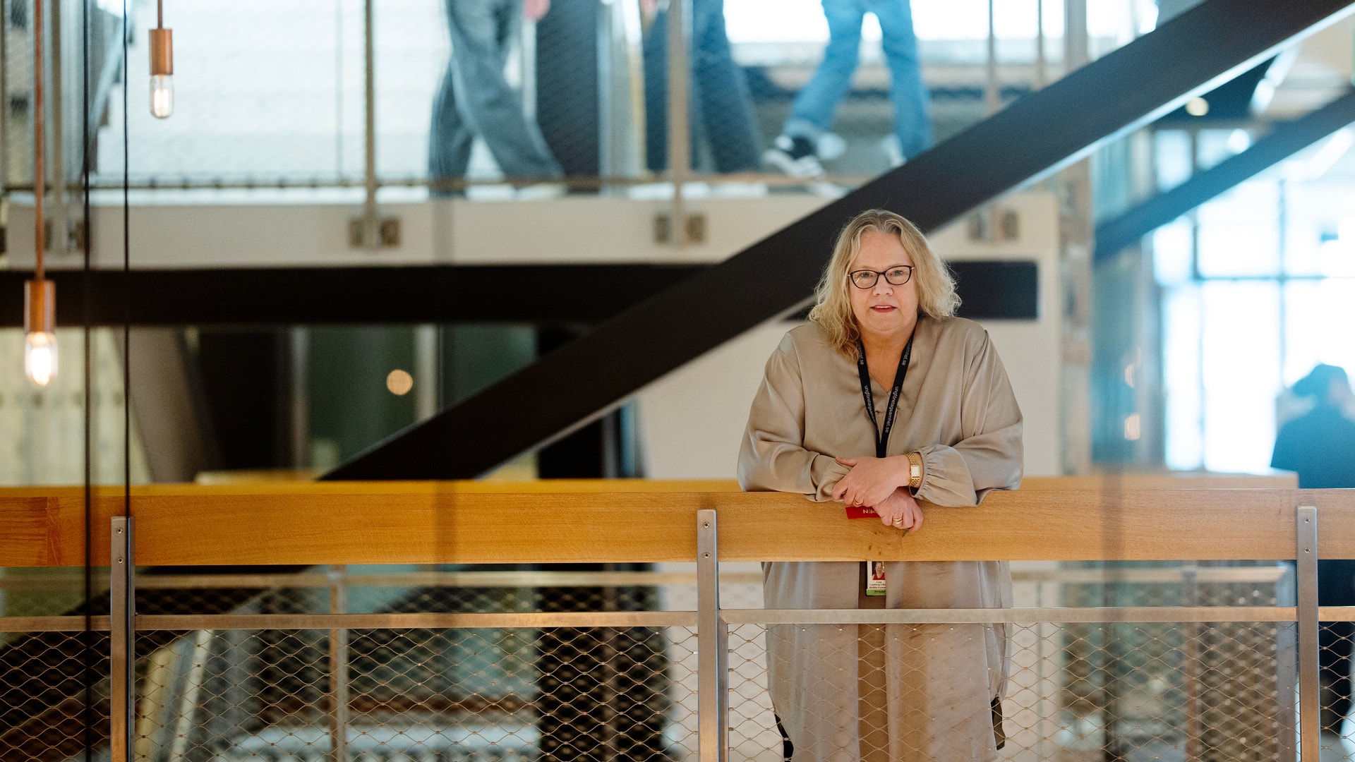 Porträtt av en kvinna med glasögon som står vid ett räcke i en modern byggnad med stora fönster och trappor i bakgrunden.