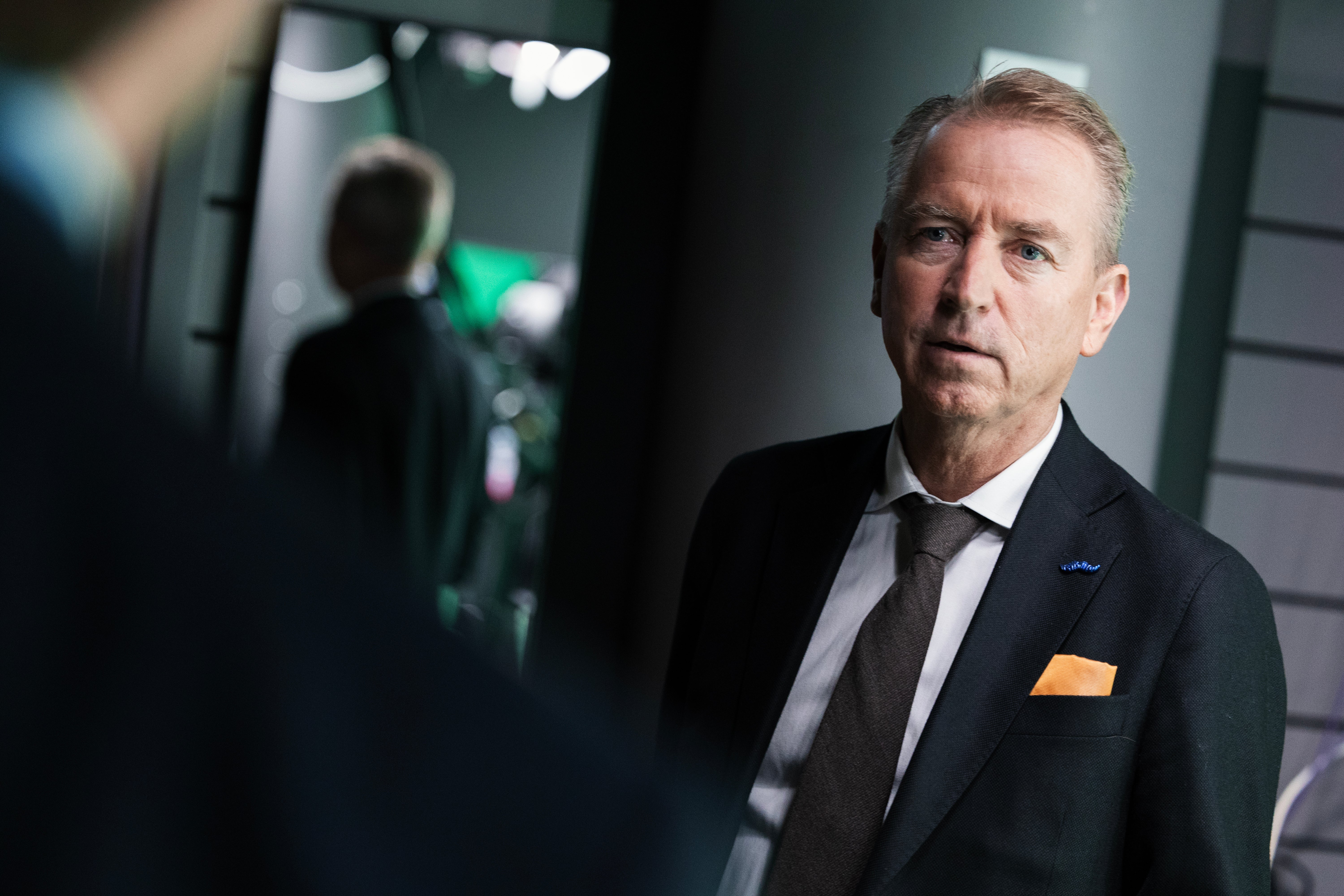 En man i mörk kostym, vit skjorta och brun slips tittar allvarligt åt sidan i en studiomiljö.