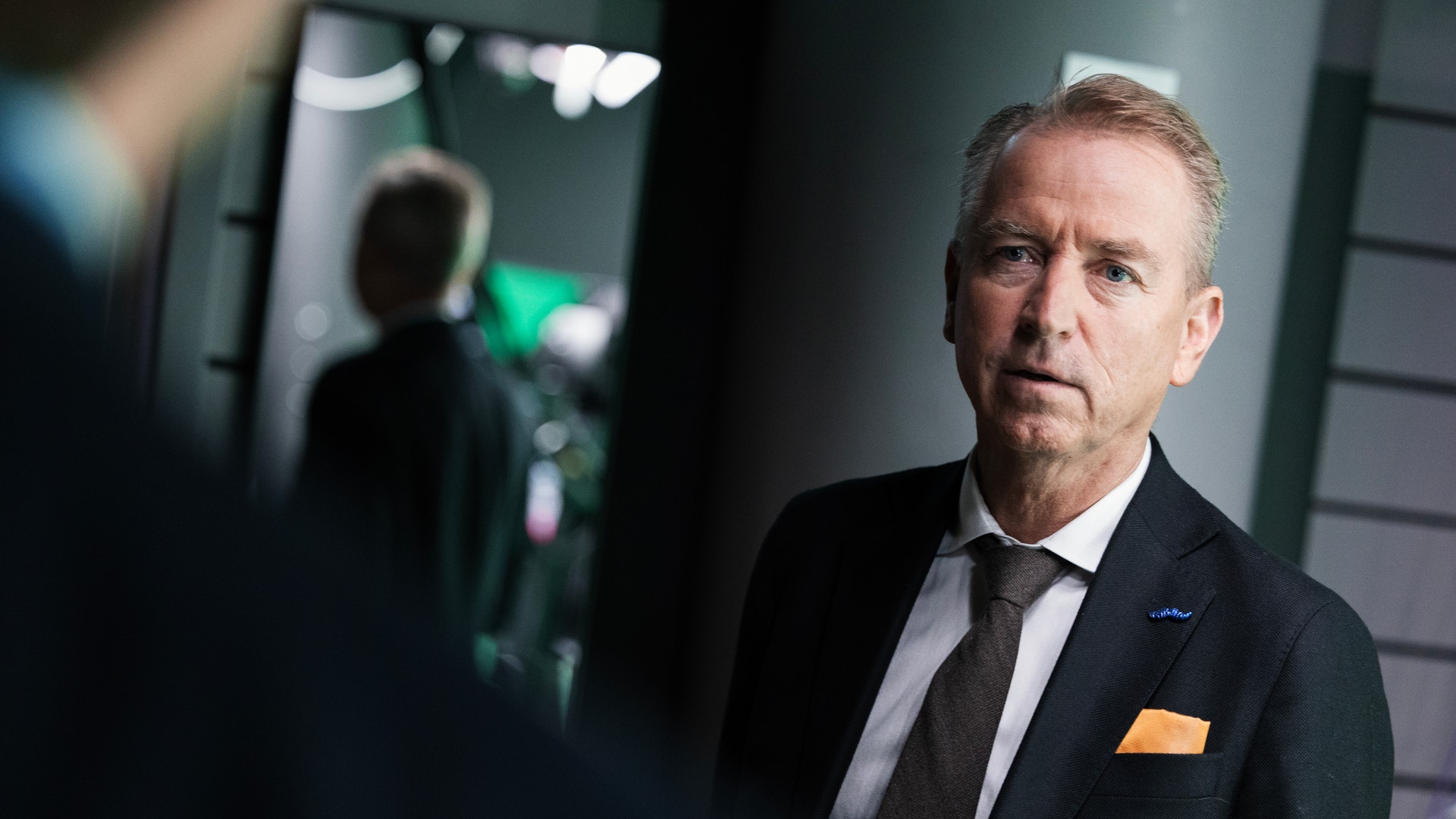 En man i mörk kostym, vit skjorta och brun slips tittar allvarligt åt sidan i en studiomiljö.