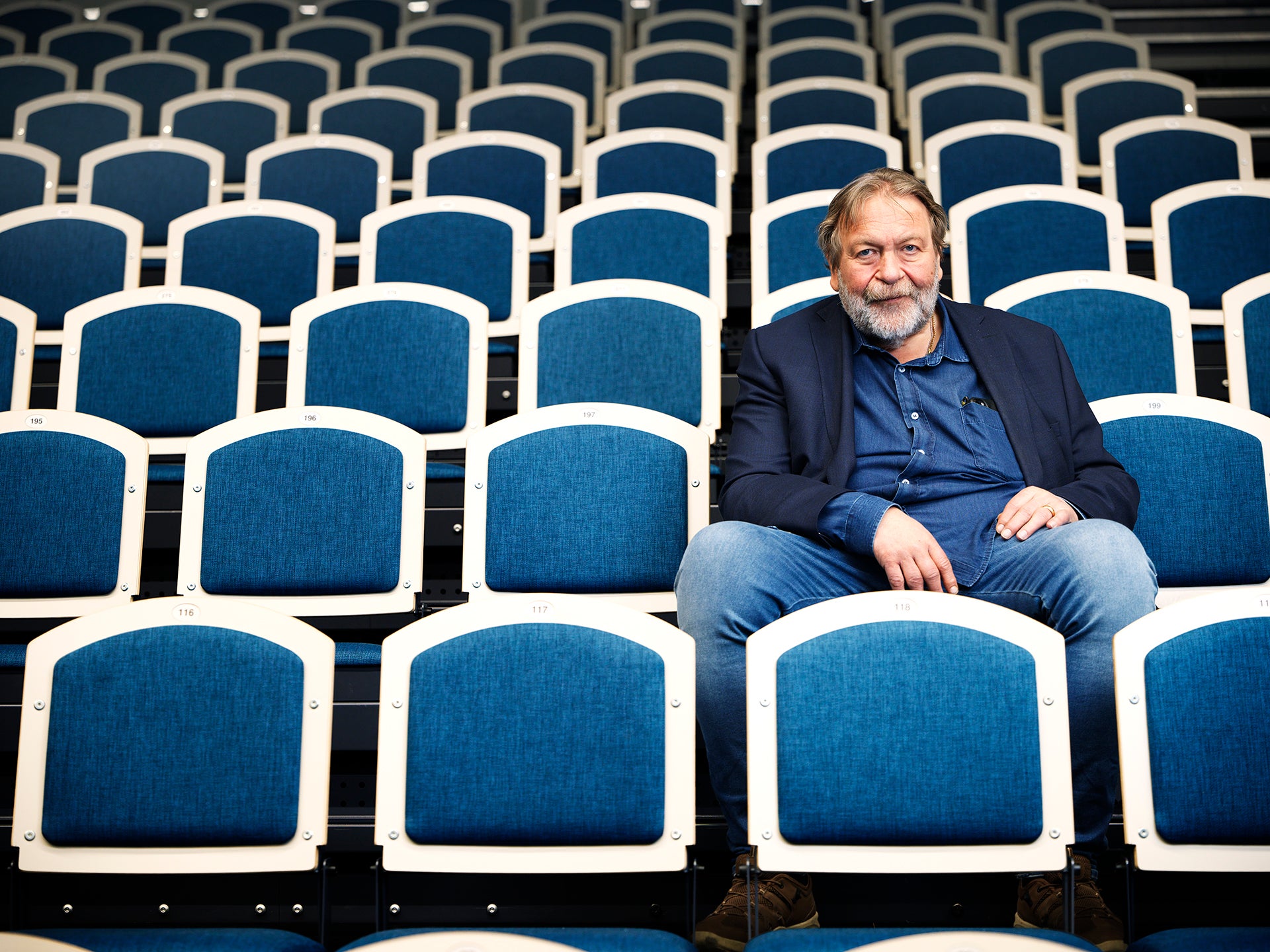En man sitter ensam bland många tomma teatersäten. Han är klädd i marinblå blazer och blå skjorta och tittar mot kameran.
