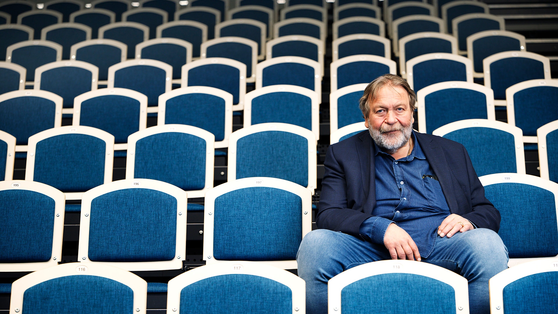 En man sitter ensam bland många tomma teatersäten. Han är klädd i marinblå blazer och blå skjorta och tittar mot kameran.
