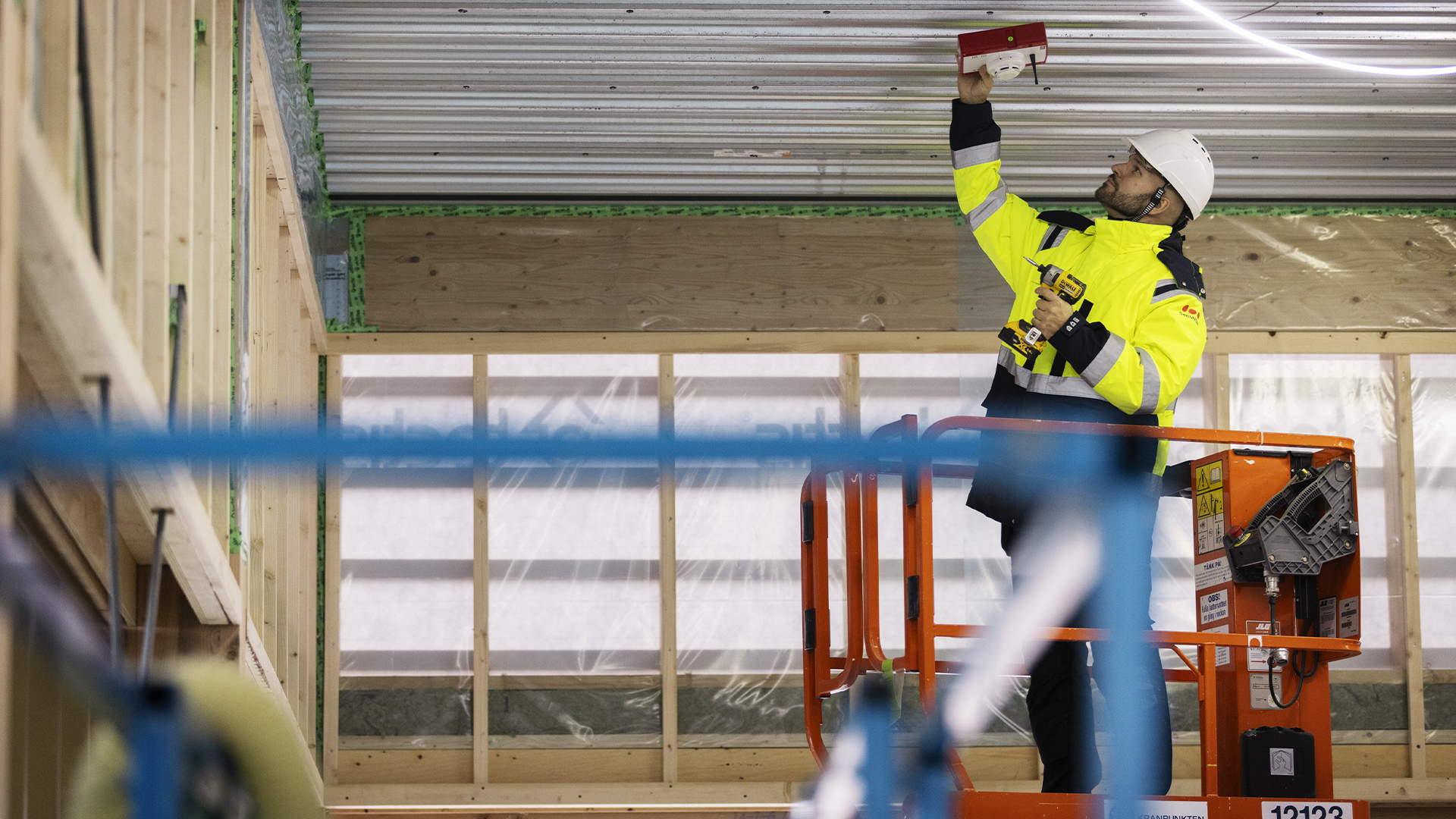 Arbetare i gul säkerhetsvest och hjälm arbetar på en orange arbetshöjdare inuti en industribyggnad och utför arbete på taket.