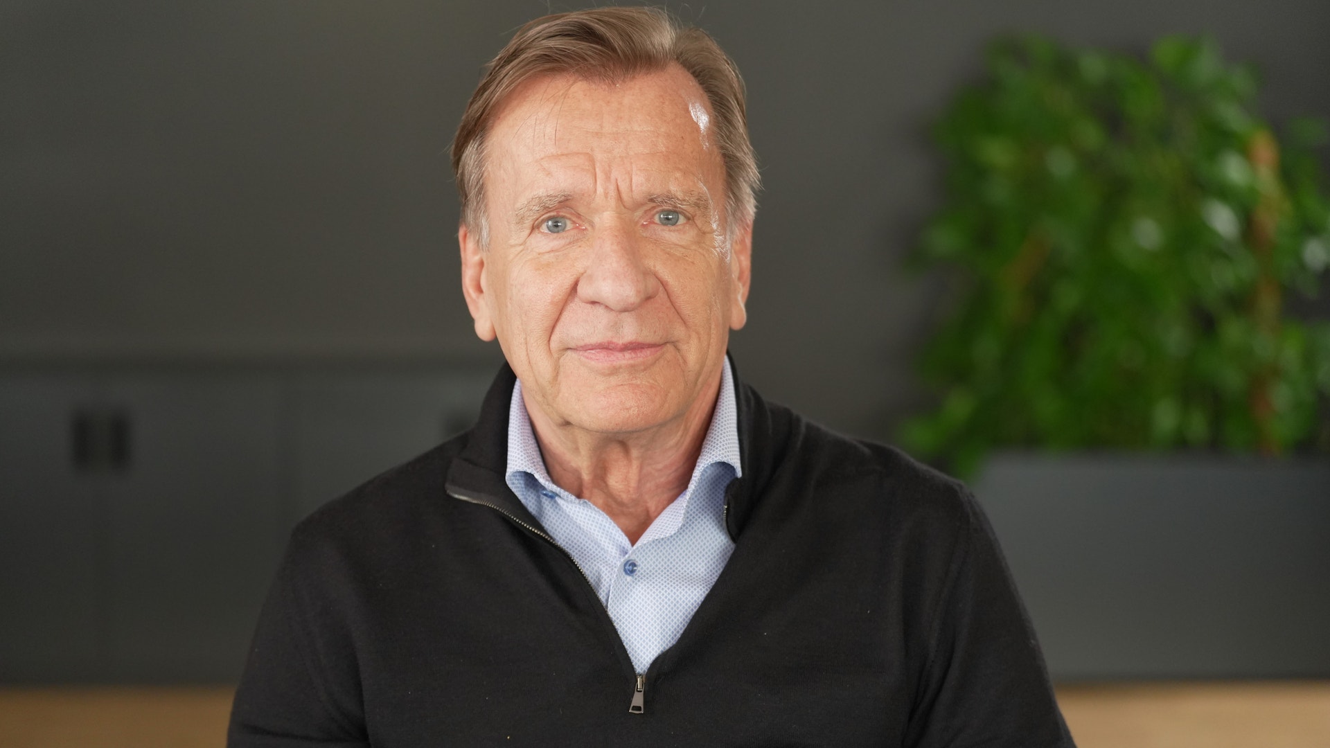 Porträtt av en äldre man i svart tröja och ljusblå skjorta mot neutral bakgrund