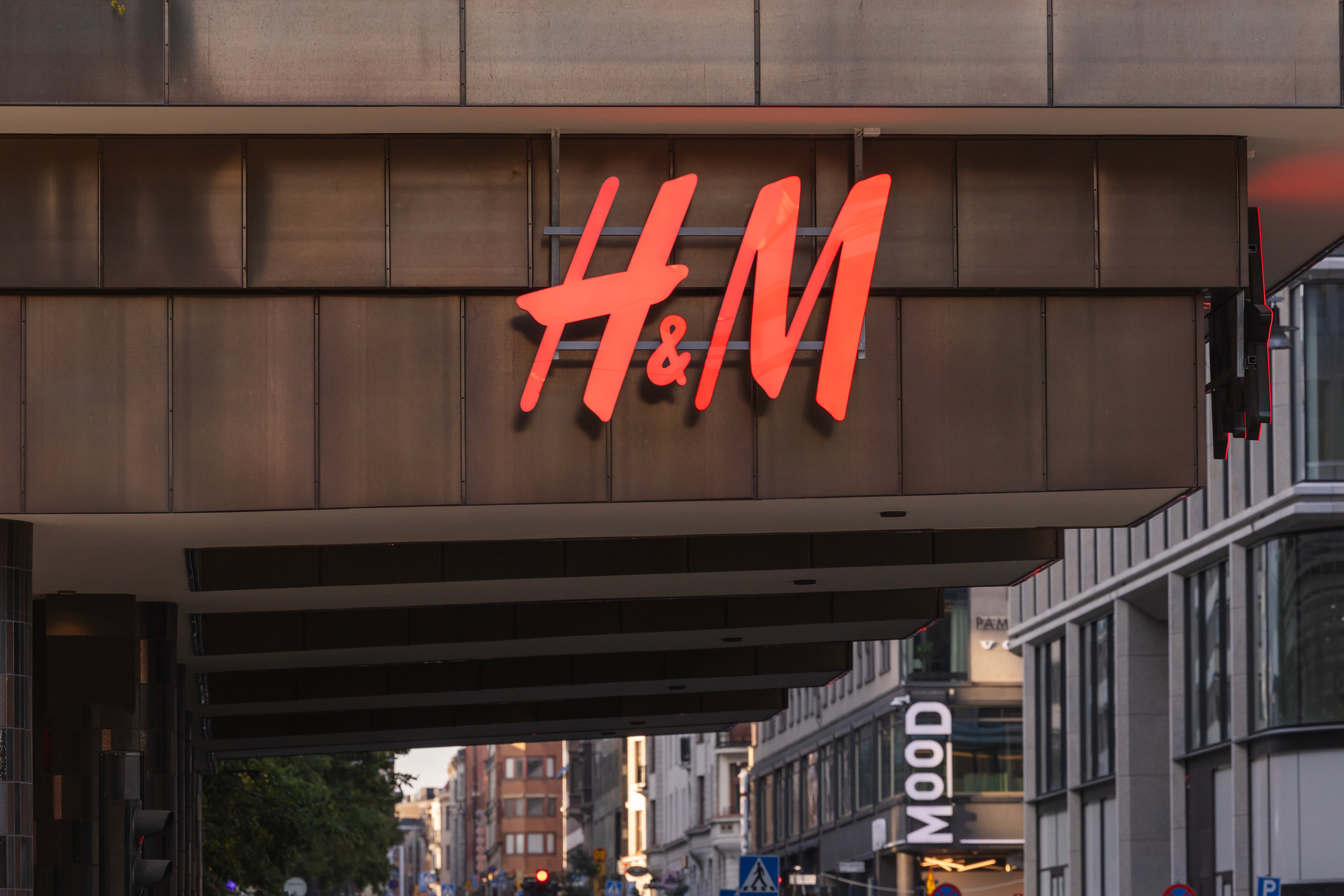 H&M:s röda neonskyllt på en brun fasad med butiksentré och stadsmiljö i bakgrunden.