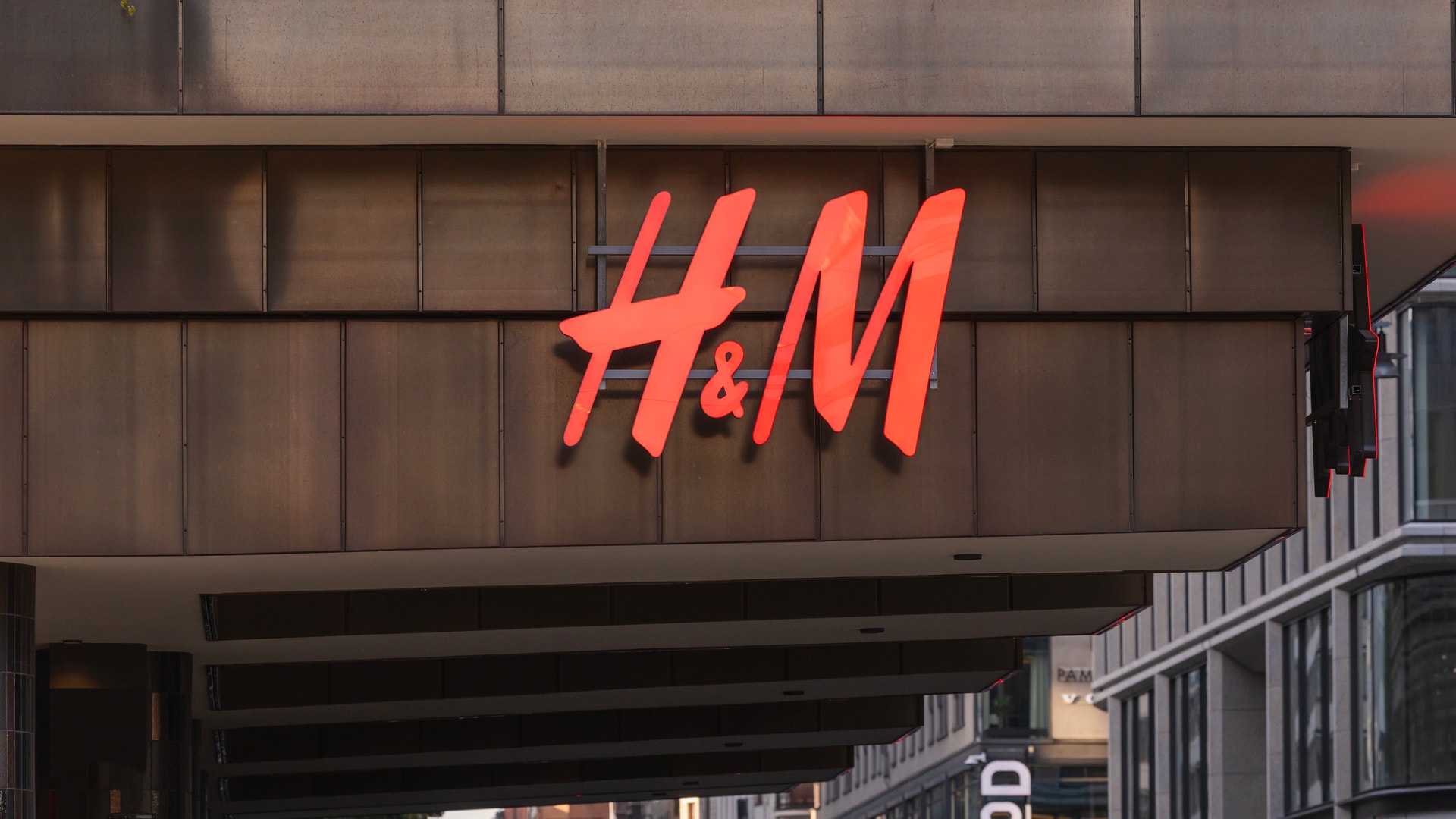 H&M:s röda neonskyllt på en brun fasad med butiksentré och stadsmiljö i bakgrunden.