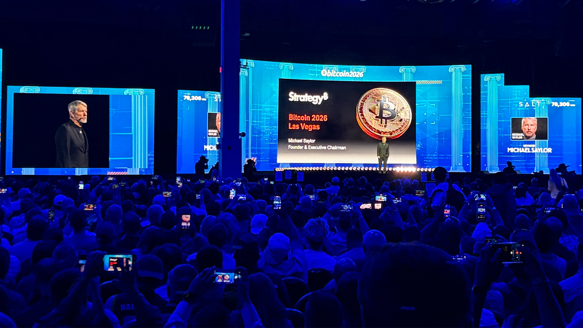 En stor konferensscen med blå belysning där en presentatör står på scenen. Stora skärmar visar presentationen med Bitcoin-tema. Publiken i förgrunden är tätt packad och många filmar med sina mobiltelefoner.