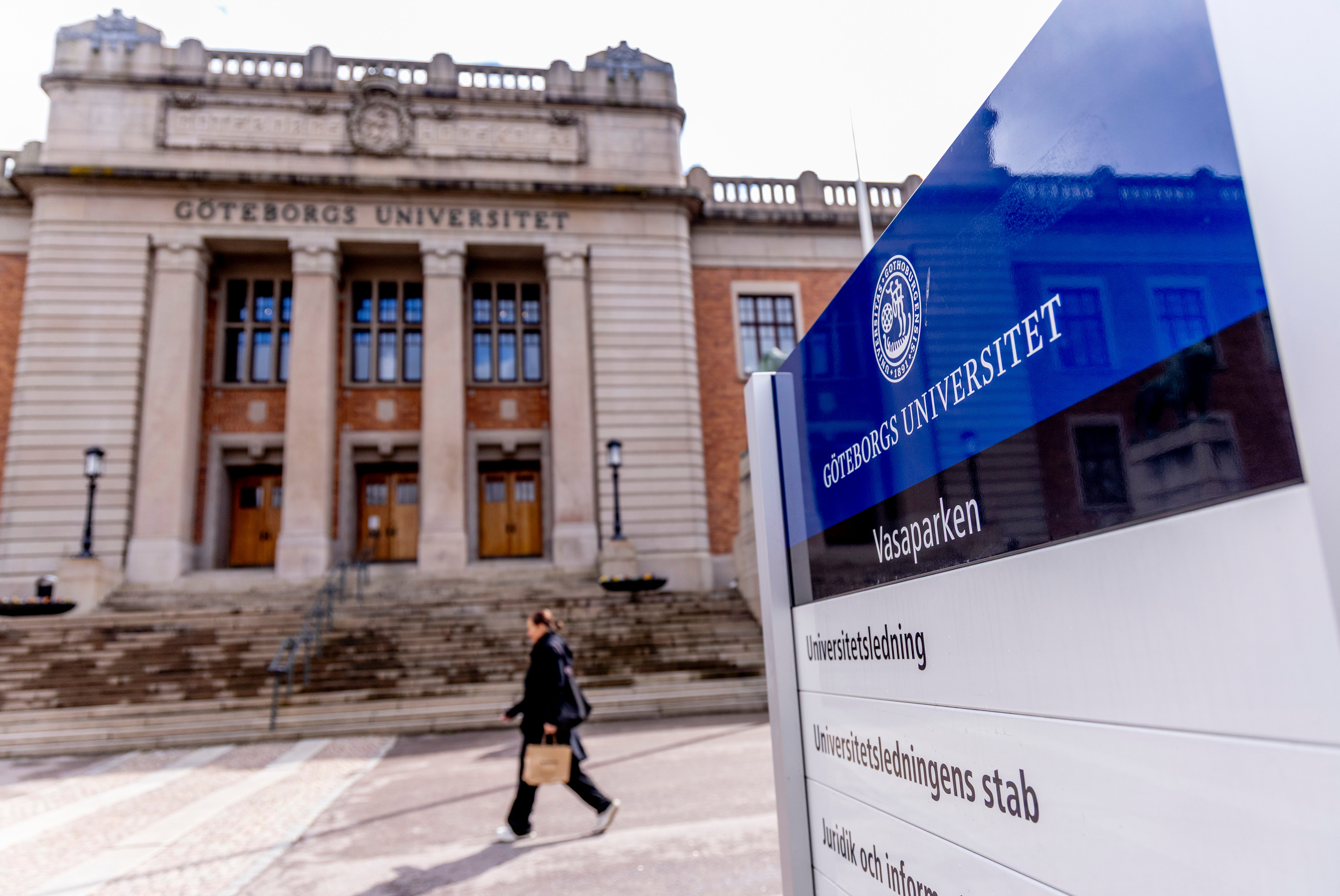 Göteborgs Universitet campus med en klassisk universitetsbyggnad och en blå informationsskylt. En person går på vägen framför byggnaden.