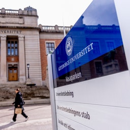 Göteborgs Universitet campus med en klassisk universitetsbyggnad och en blå informationsskylt. En person går på vägen framför byggnaden.
