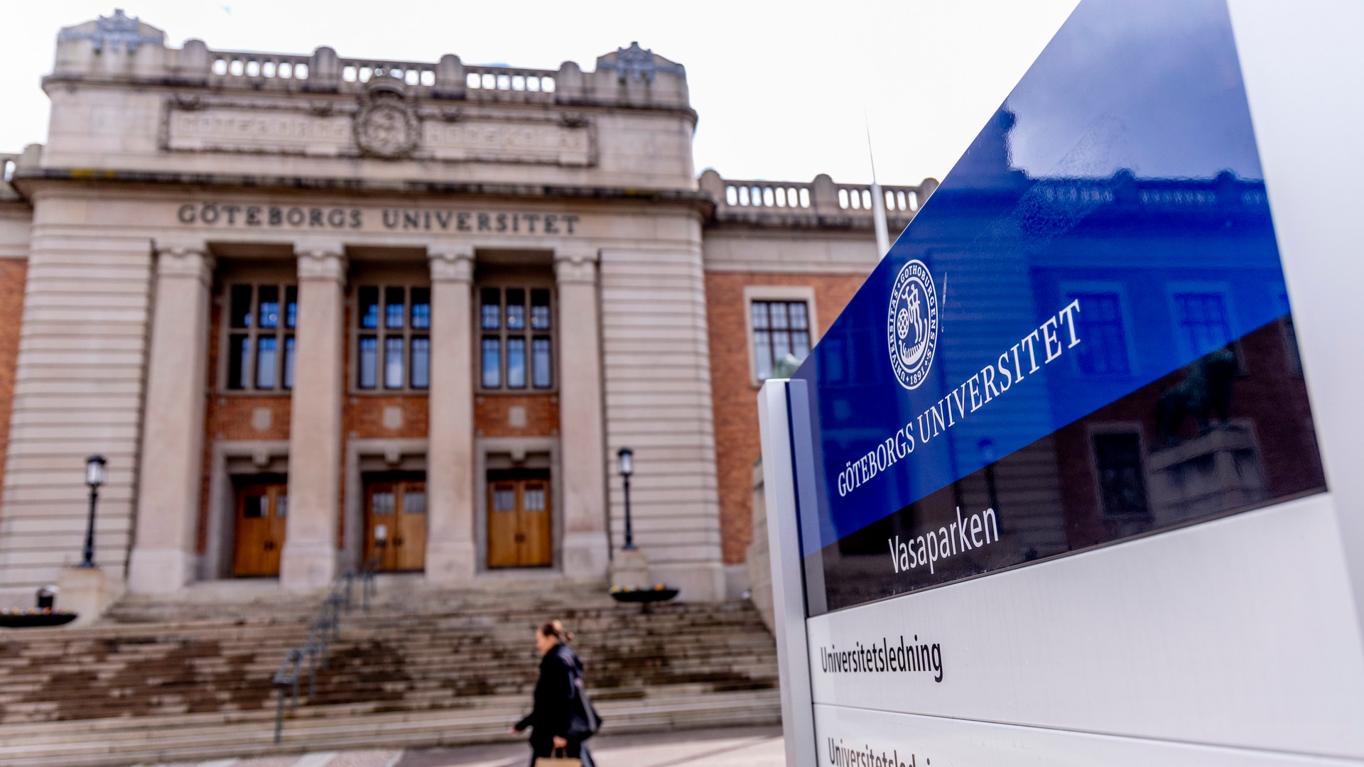 Göteborgs Universitet campus med en klassisk universitetsbyggnad och en blå informationsskylt. En person går på vägen framför byggnaden.