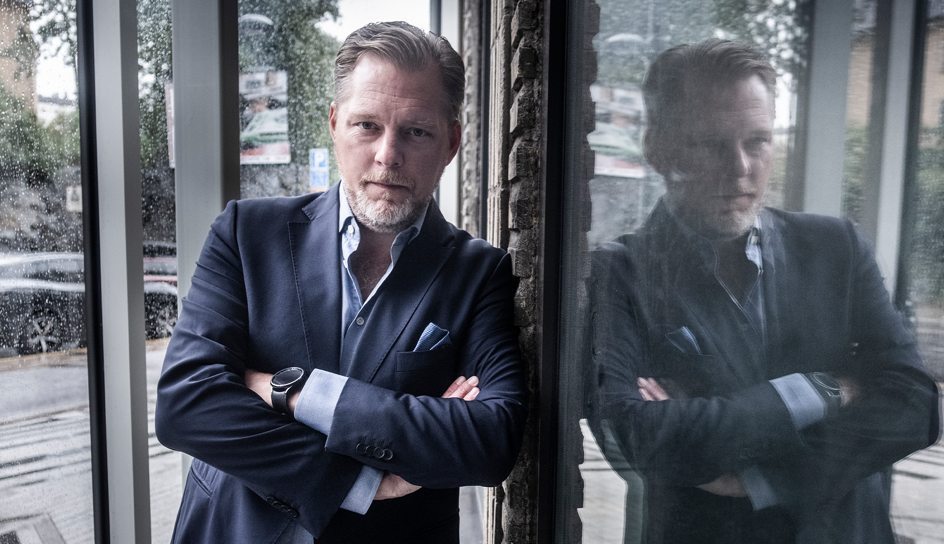 Man i mörk kostym står vid ett fönster med armarna i kors. Hans spegelbild syns i glaset bakom honom.