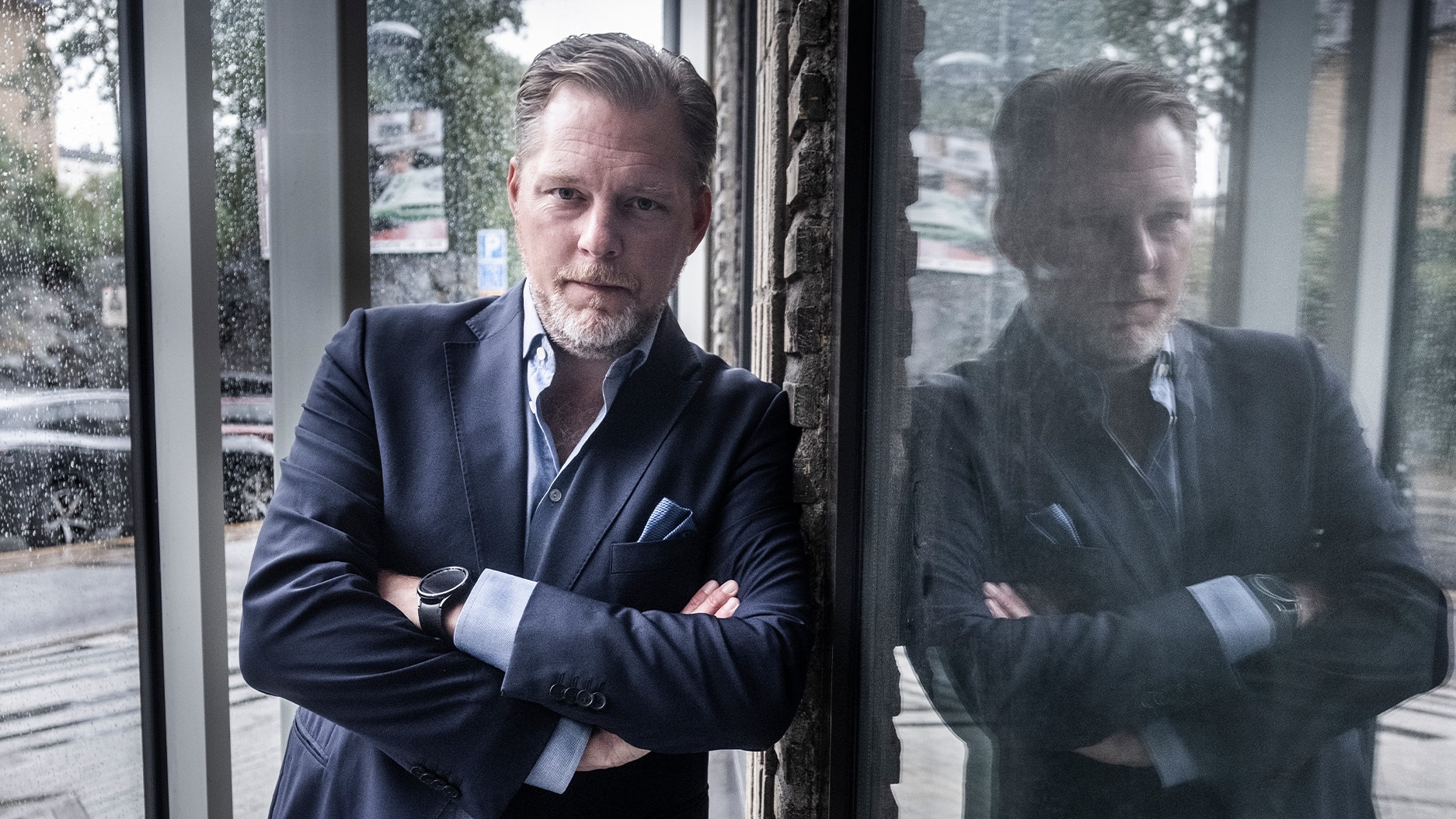 Man i mörk kostym står vid ett fönster med armarna i kors. Hans spegelbild syns i glaset bakom honom.
