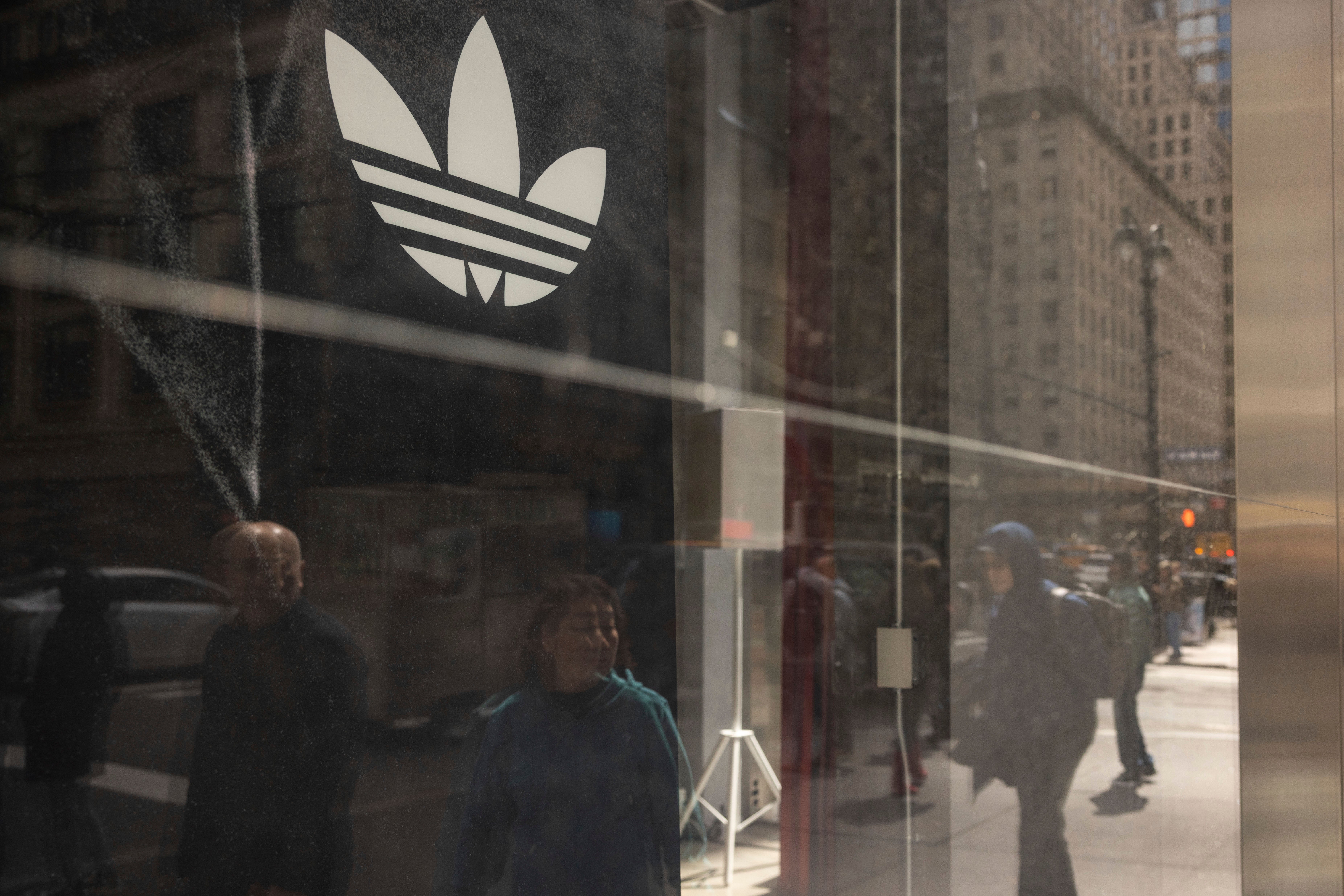 Adidas-butikens skyltfönster med det vita Adidas-logotypen på glas, med gatuscenen och människor reflekterade i fönstret.
