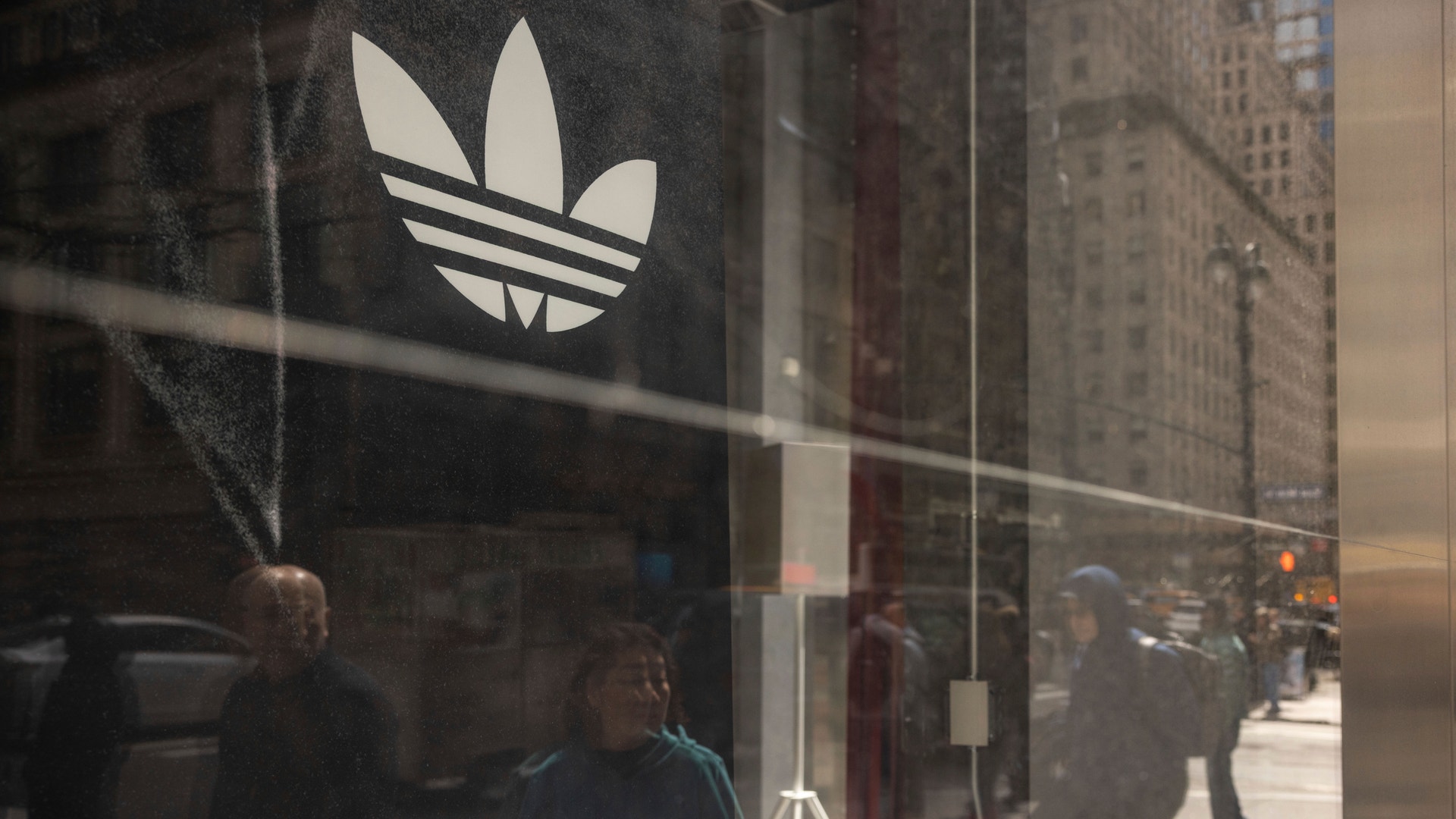 Adidas-butikens skyltfönster med det vita Adidas-logotypen på glas, med gatuscenen och människor reflekterade i fönstret.
