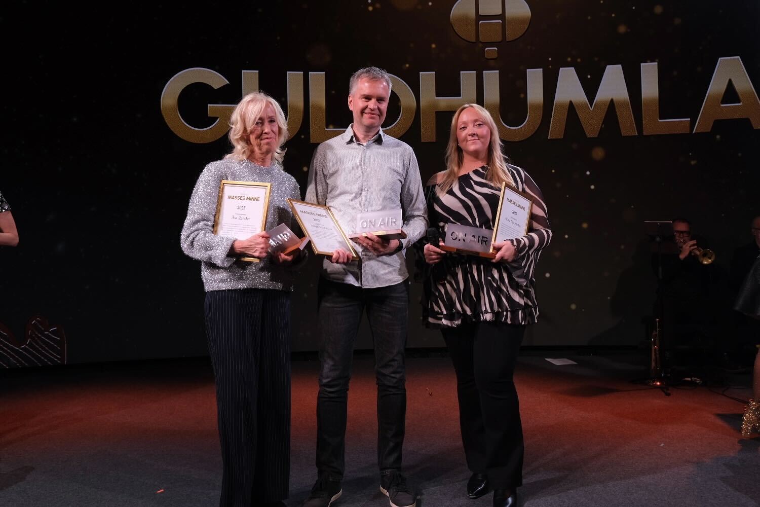 Tre personer står på en scen under en ceremoni och håller upp diplom eller utmärkelser. I bakgrunden syns texten Guldhumla.