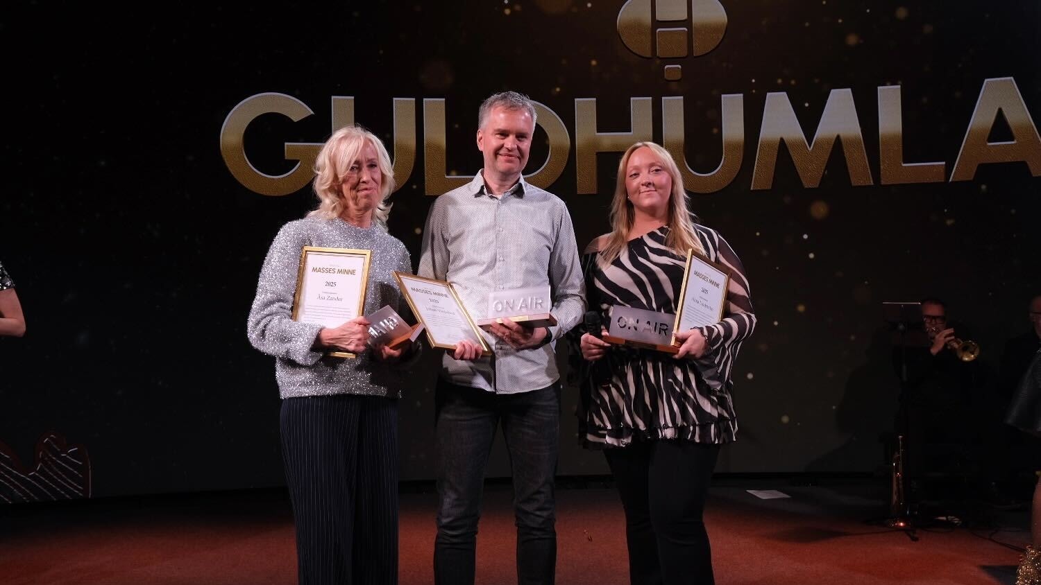 Tre personer står på en scen under en ceremoni och håller upp diplom eller utmärkelser. I bakgrunden syns texten Guldhumla.