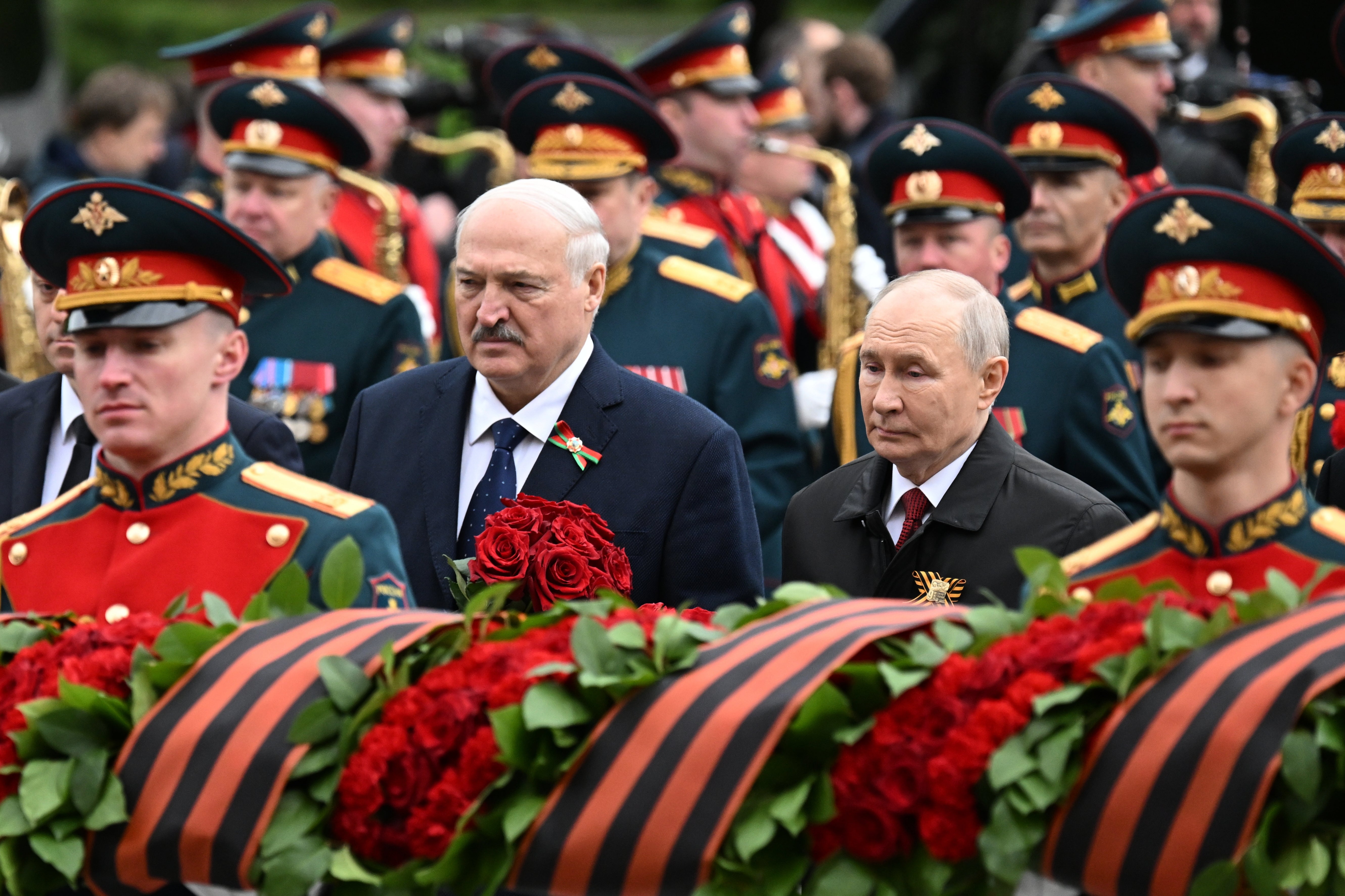 Militär ceremoni med uniformerade officerare och två civila män vid en minnesstund. Röda rosor och svart-orangea band är synliga i förgrunden.
