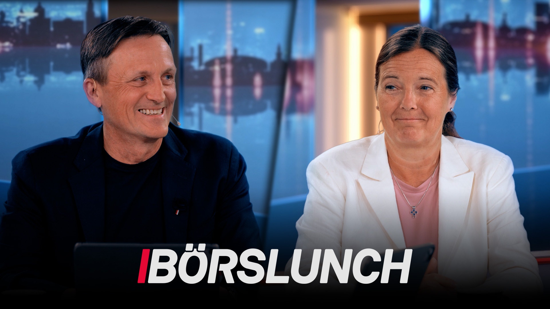 En man och en kvinna sitter och samtalar framför ett fönster med stadsvy. Texten BÖRSLUNCH visas längst ner på bilden.