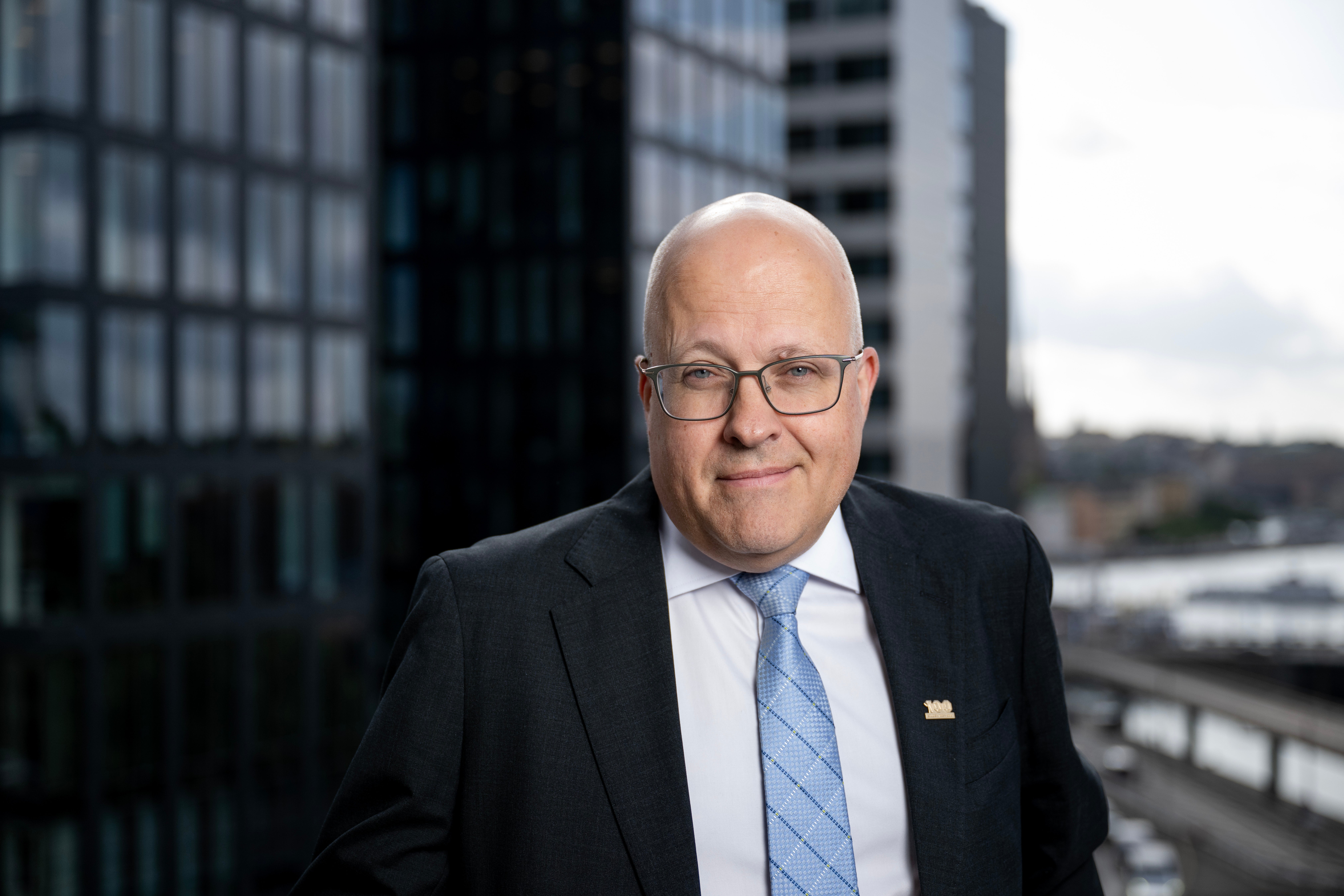Porträtt av en äldre man med glasögon och kostym framför moderna kontorsbyggnader