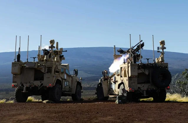 Två bepansrade militärfordon i sandfärg står bredvid varandra på en torr slätt. Från det högra fordonet avfyras ett vapen med synlig rök och eld. I bakgrunden syns en bergssluttning under klarblå himmel.