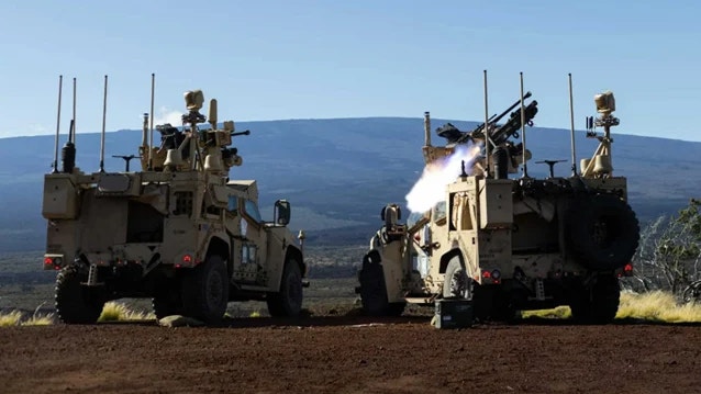 Två bepansrade militärfordon i sandfärg står bredvid varandra på en torr slätt. Från det högra fordonet avfyras ett vapen med synlig rök och eld. I bakgrunden syns en bergssluttning under klarblå himmel.