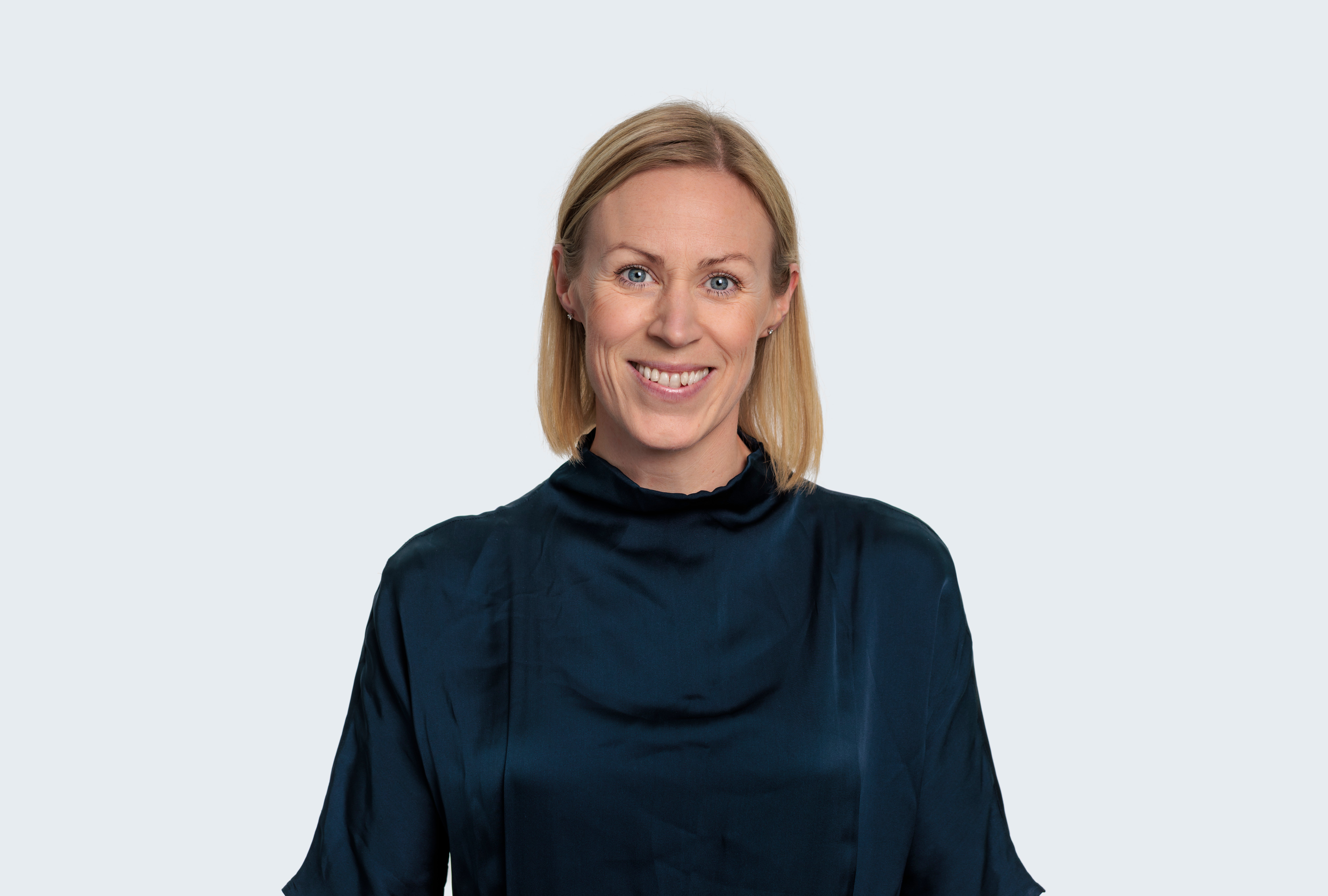 Porträtt av en leende kvinna med axellångt blont hår, klädd i en mörkblå blus, mot en ljus bakgrund.