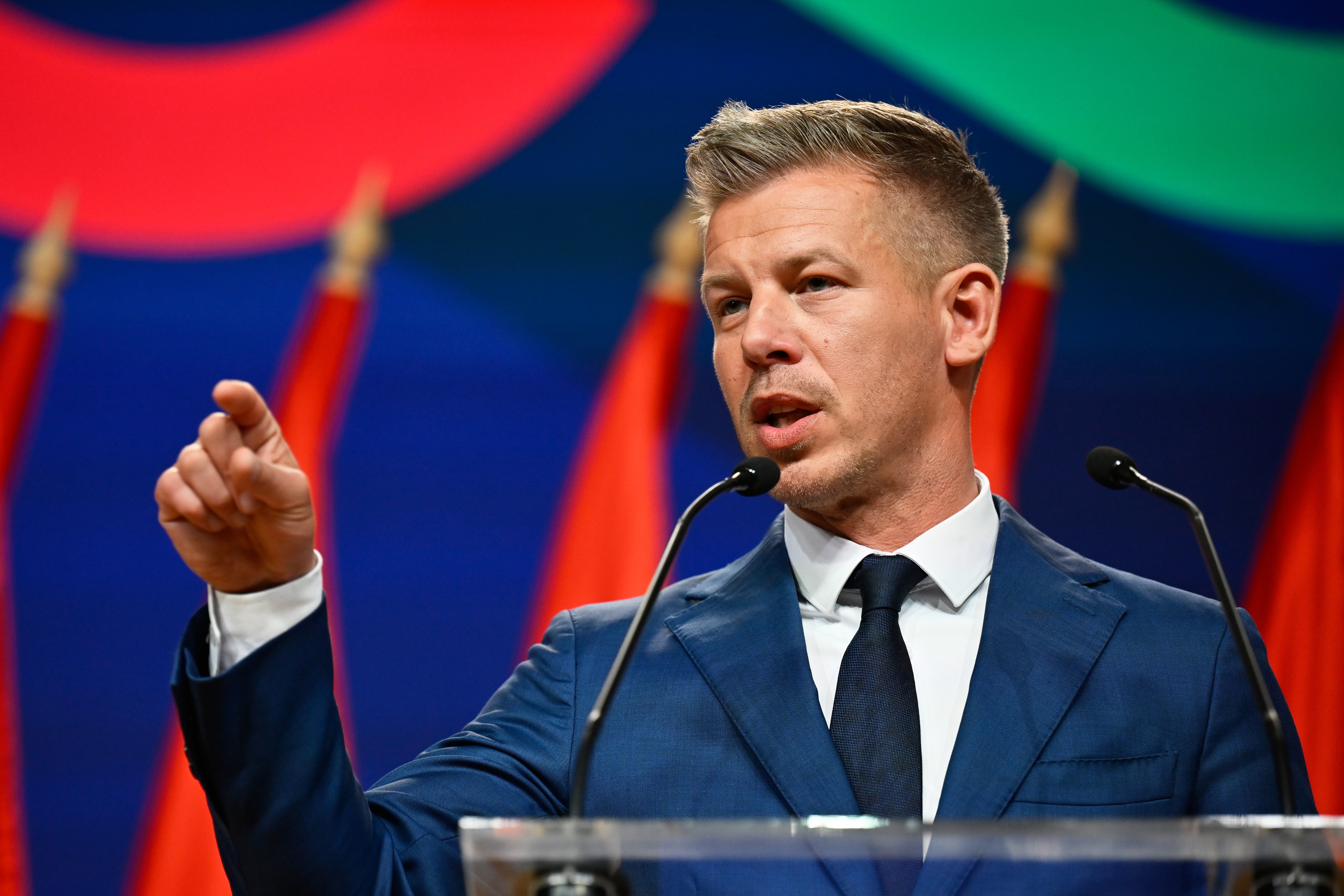 Man i mörk blå kostym talar vid ett podium med mikrofon framför en färgglad bakgrund.