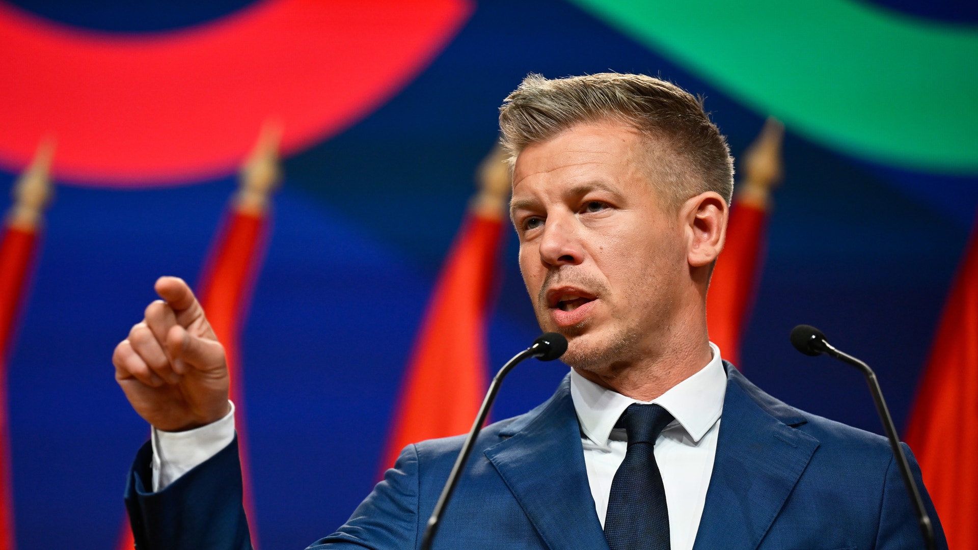 Man i mörk blå kostym talar vid ett podium med mikrofon framför en färgglad bakgrund.