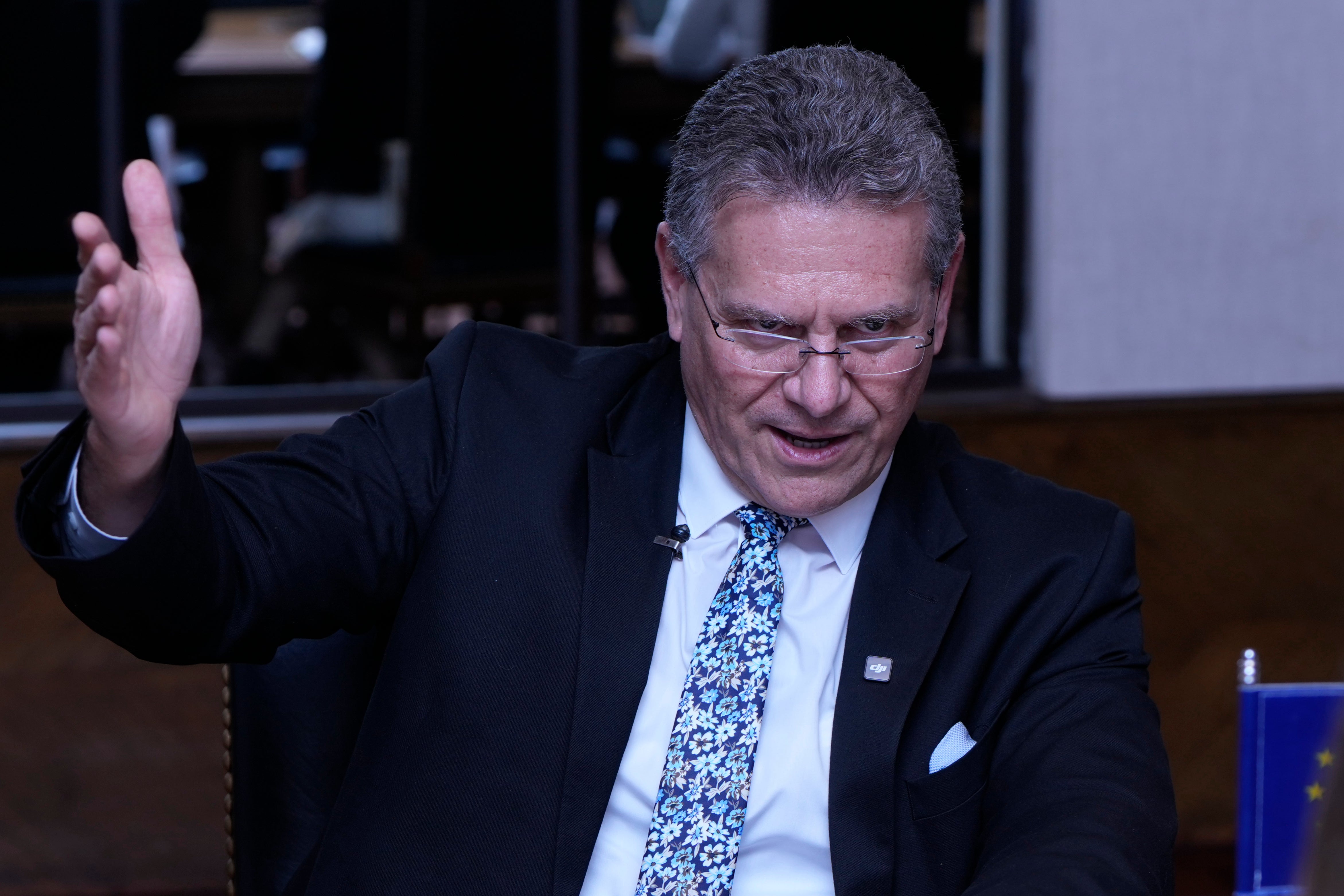 En man i mörk kostym och blå prickig slips gestikulerar medan han talar på en scen.
