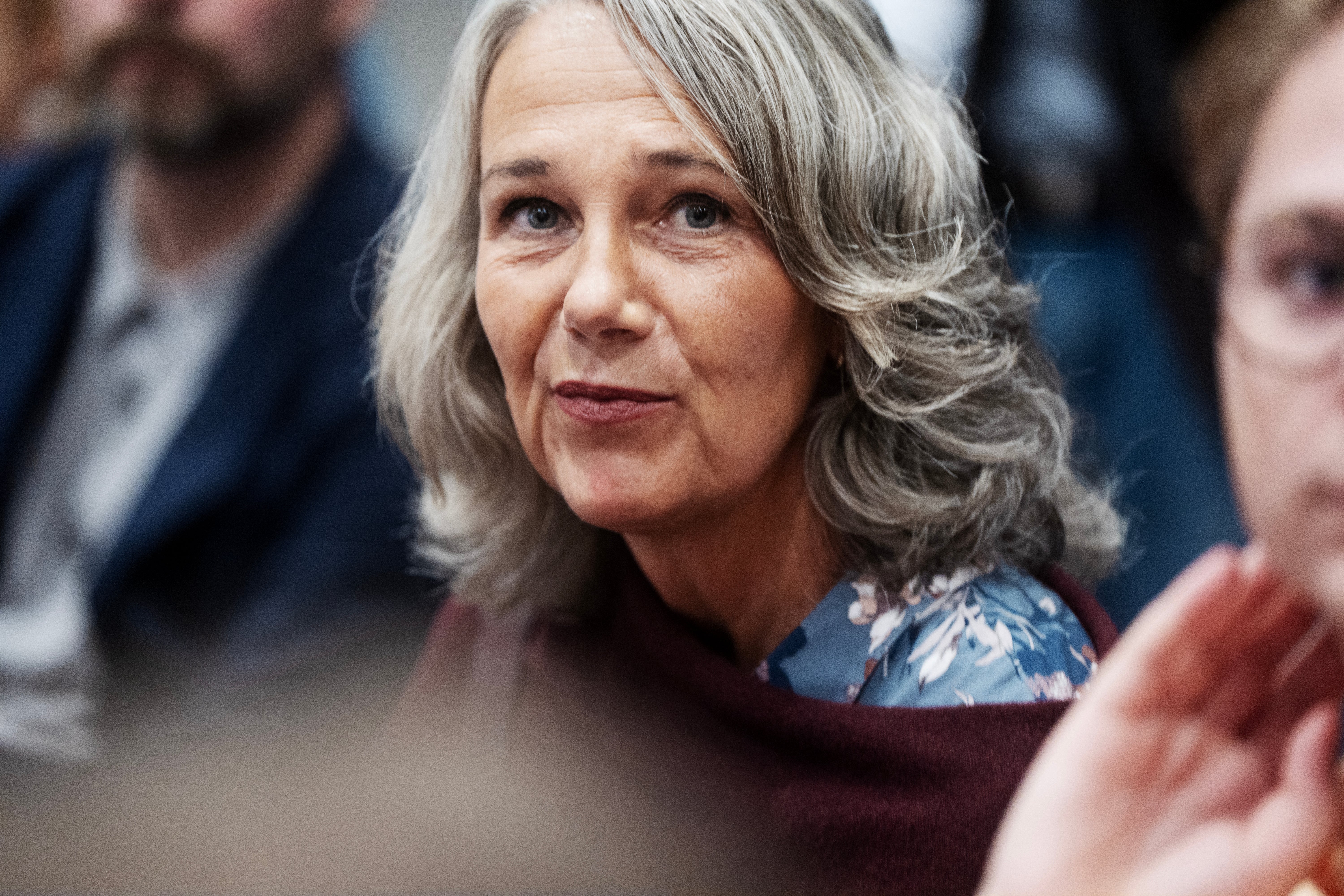 Porträtt av en äldre kvinna med långt gråvitt hår på ett evenemang med andra människor i bakgrunden.