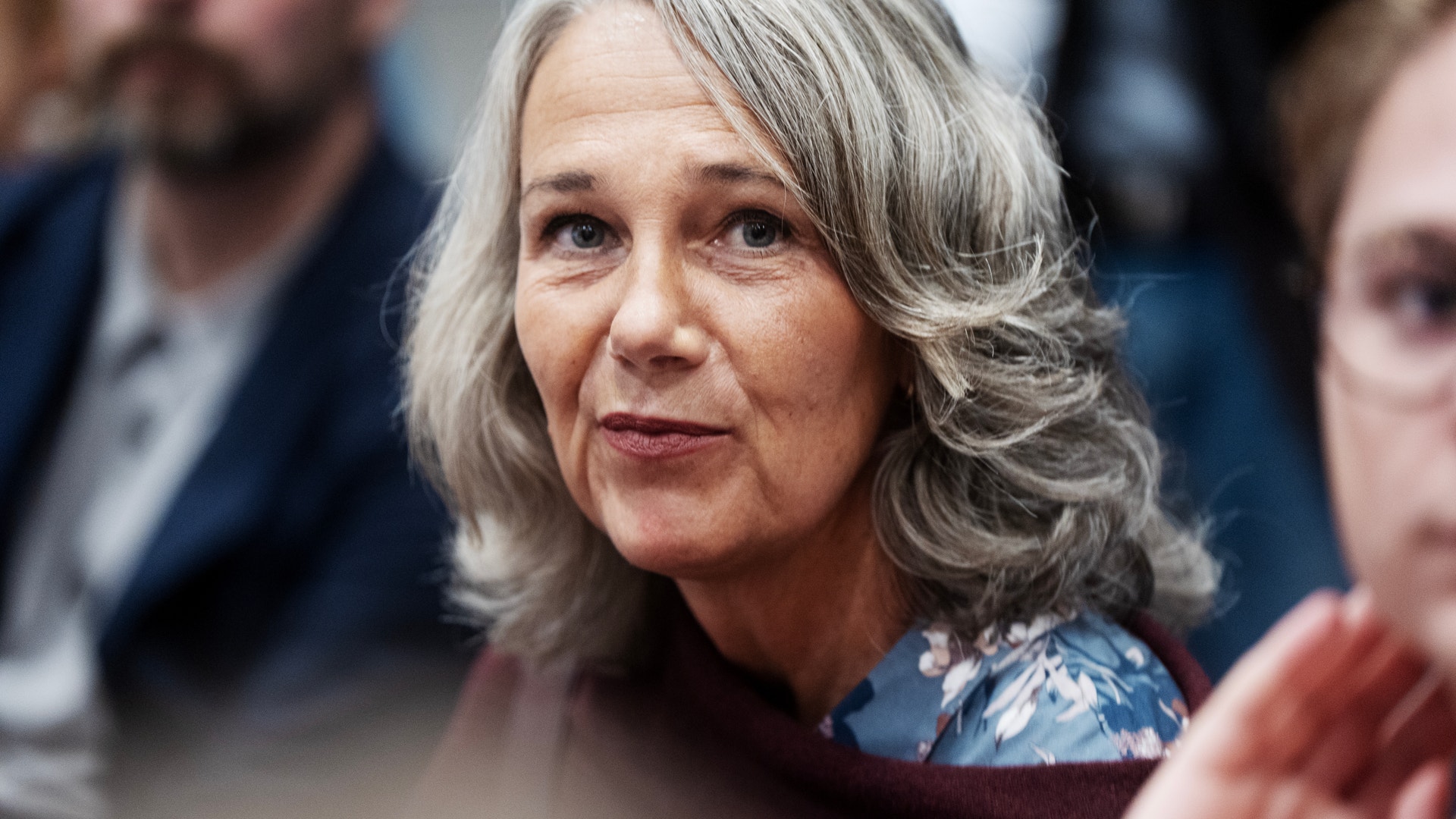 Porträtt av en äldre kvinna med långt gråvitt hår på ett evenemang med andra människor i bakgrunden.