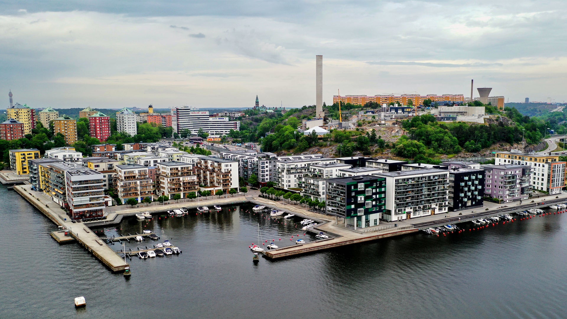 Flygfoto över Stockholm visar moderna bostadshus längs vattnet med färgglada byggnader och stadens skyline i bakgrunden.