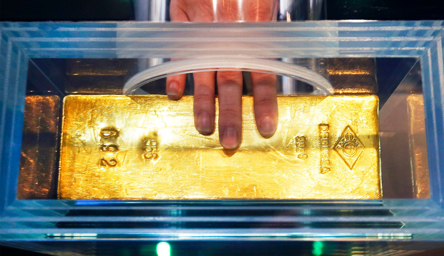 En guldtacka i en glaskupol. En hand med röd nagellack pekar på guldtackan.