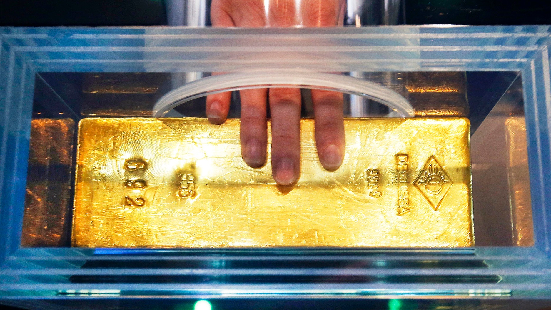 En guldtacka i en glaskupol. En hand med röd nagellack pekar på guldtackan.