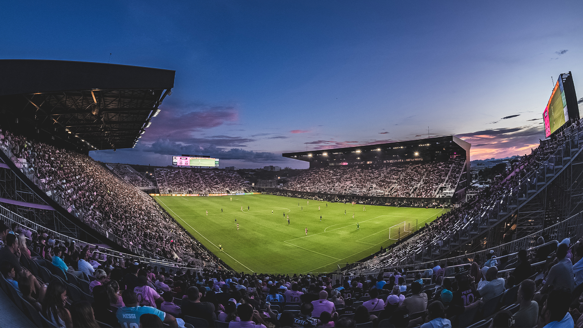En fullsatt fotbollsstadion under en match på kvällen med publik på läktarna, grönt gräs och floodlights tända.