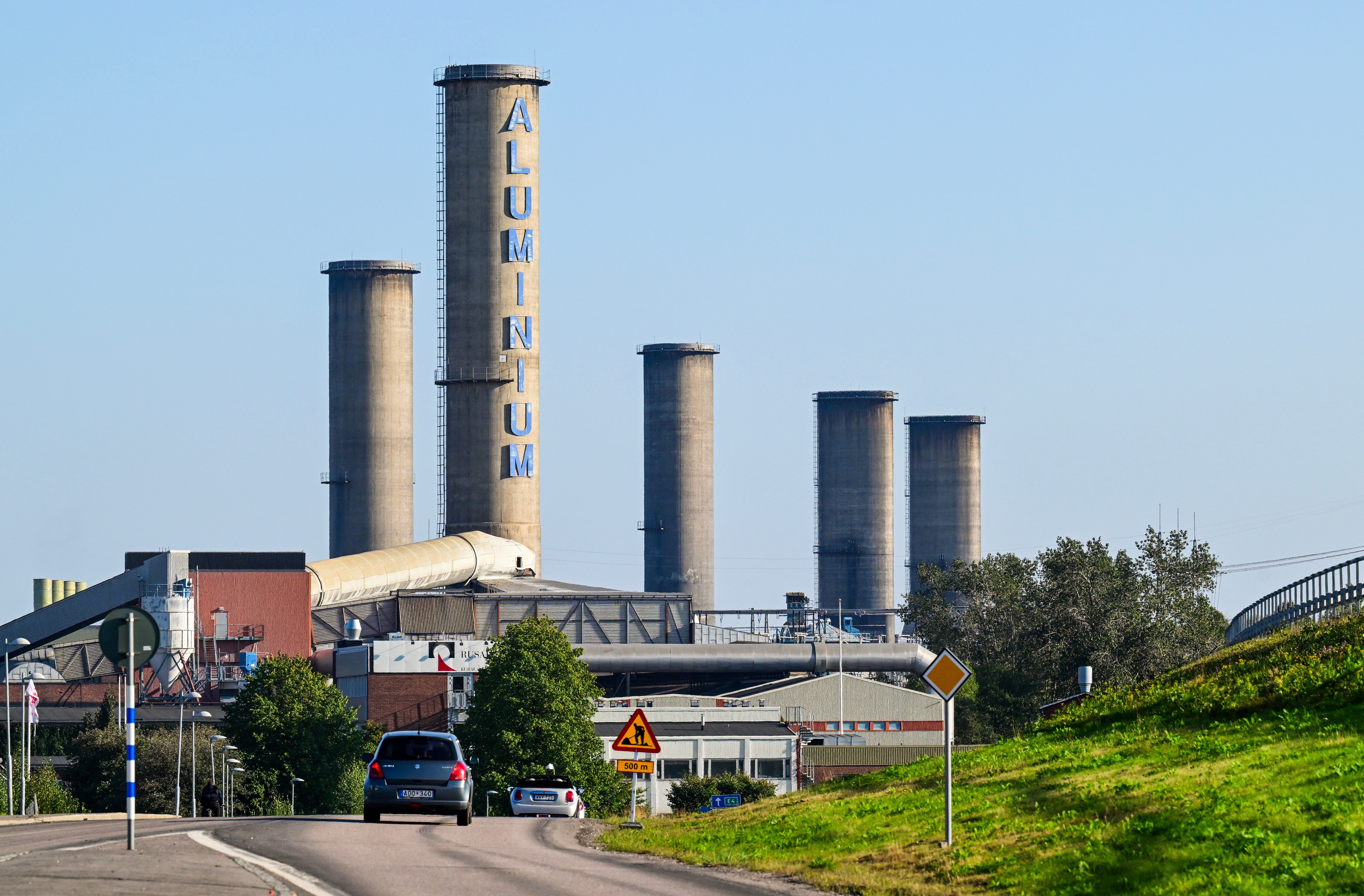 Industrianläggning med flera höga cementrör och röda byggnader vid en väg.