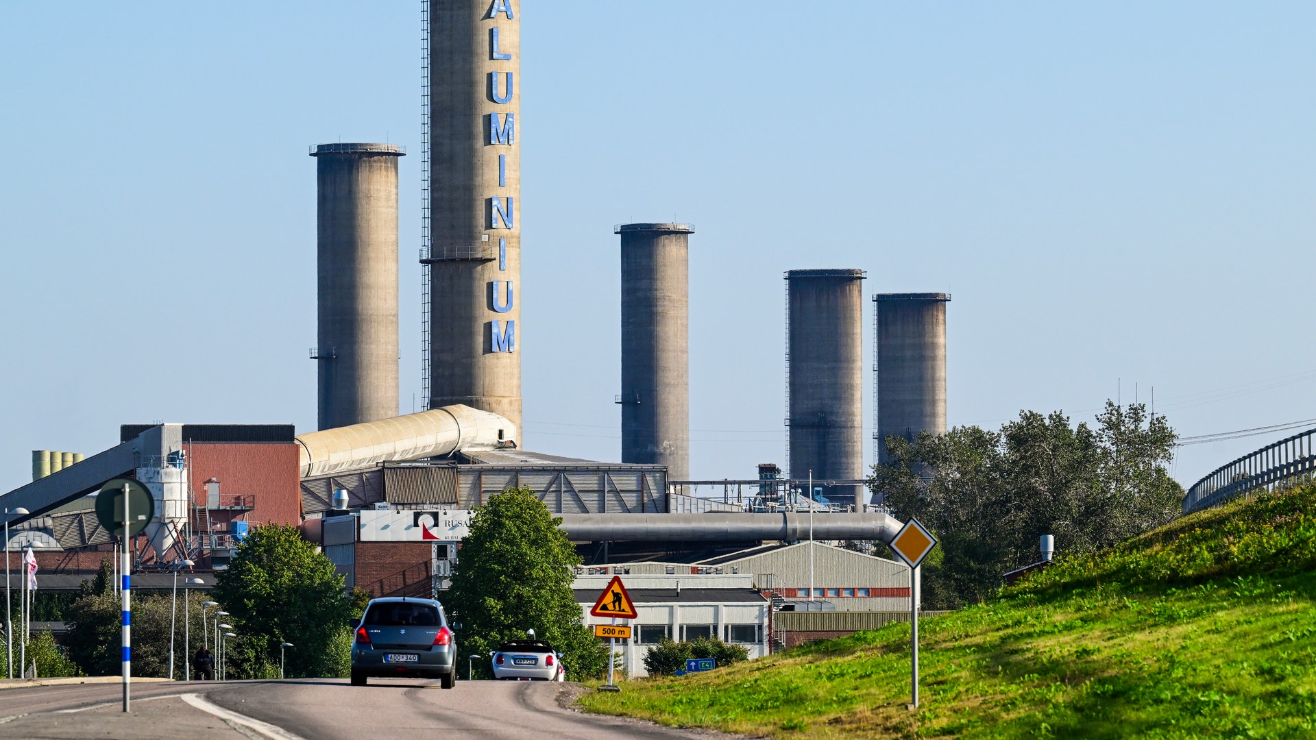Industrianläggning med flera höga cementrör och röda byggnader vid en väg.