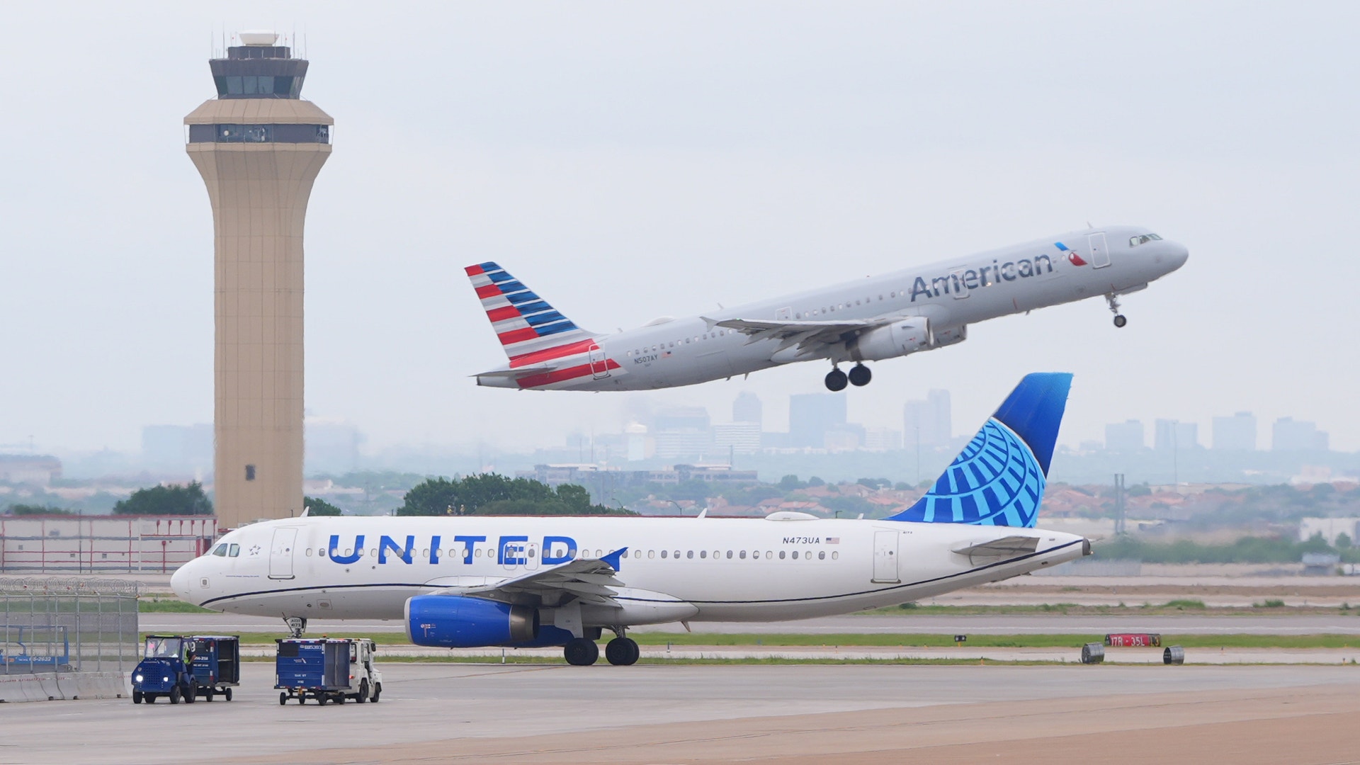 Två flygplan på en flygplats med ett kontrolltorn i bakgrunden. Ett United-plan står på marken medan ett American Airlines-plan lyfter.