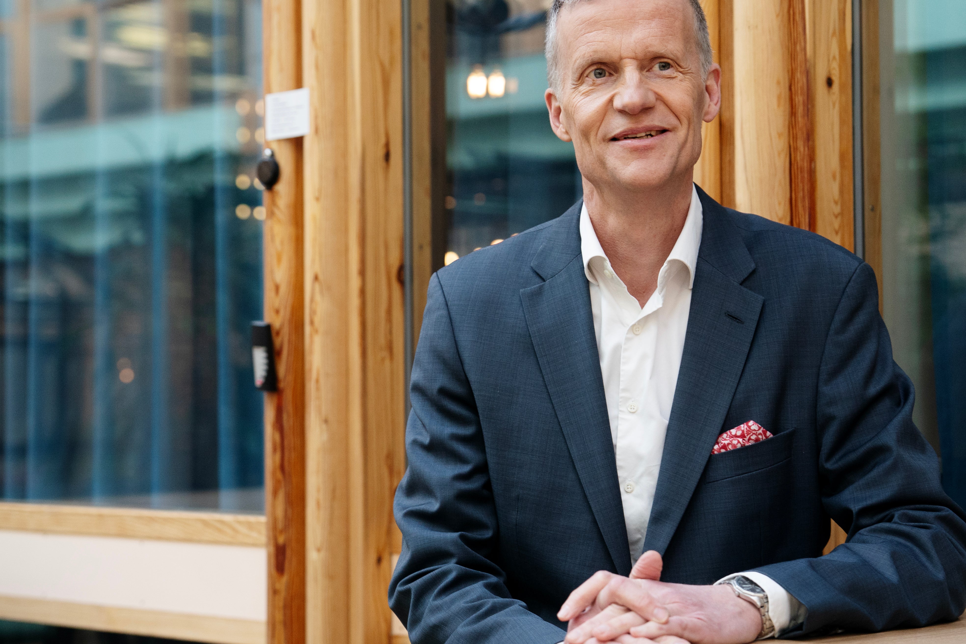 Porträtt av en man i mörkblå kostym och vit skjorta framför en trävägg med glasfönster i bakgrunden.